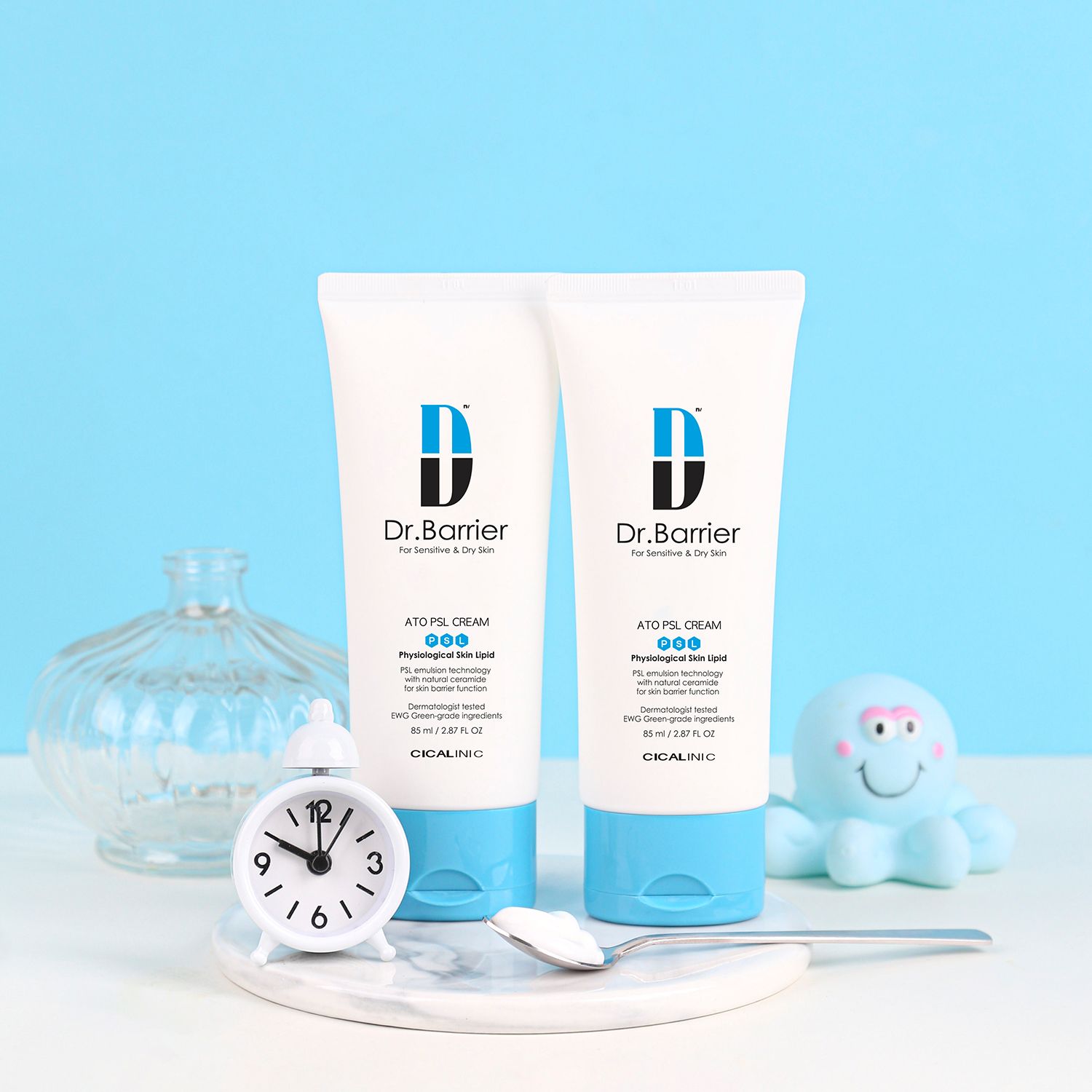 Due tubi di Dr.Barrier ATO PSL Cream. Su una superficie con orologio, cucchiaio e decorazioni. Schema colori blu e bianco.