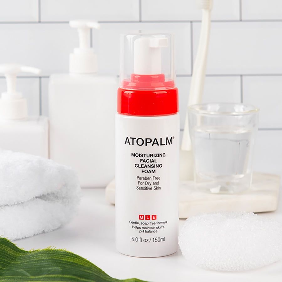 Flacone ATOPALM Moisturizing Facial Cleansing Foam, bianco con coperchio rosso. Su tavolo con accessori.