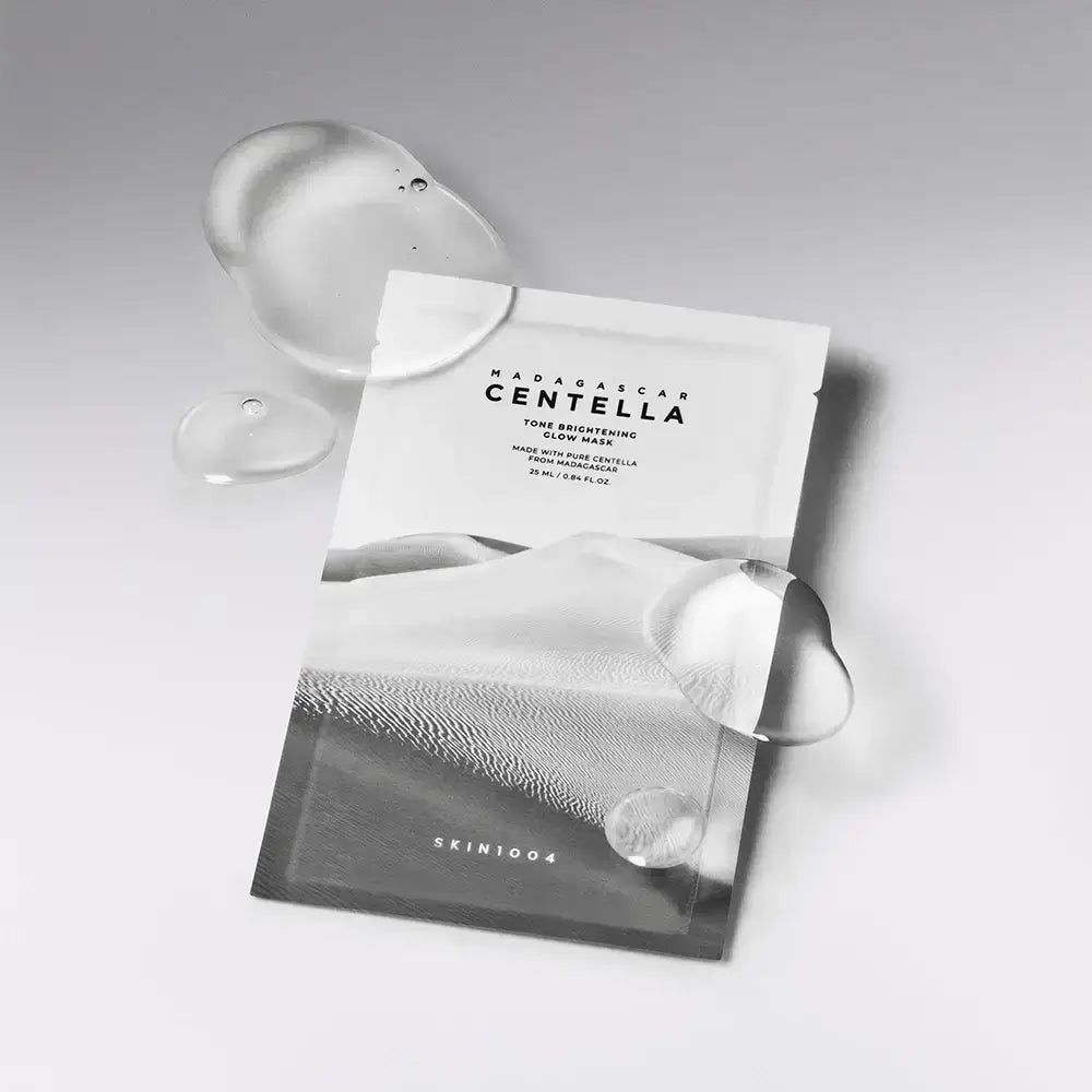 Confezione della maschera SKIN1004 Glow. Bustina bianca con stampa paesaggistica e nome del prodotto. Gocce di liquido trasparente accanto.