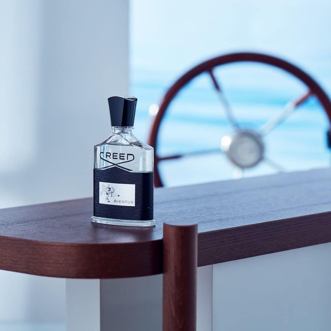 Flacone in vetro con tappo nero. Scritta "CREED". Etichetta nera con "AVENTUS". Su superficie in legno.