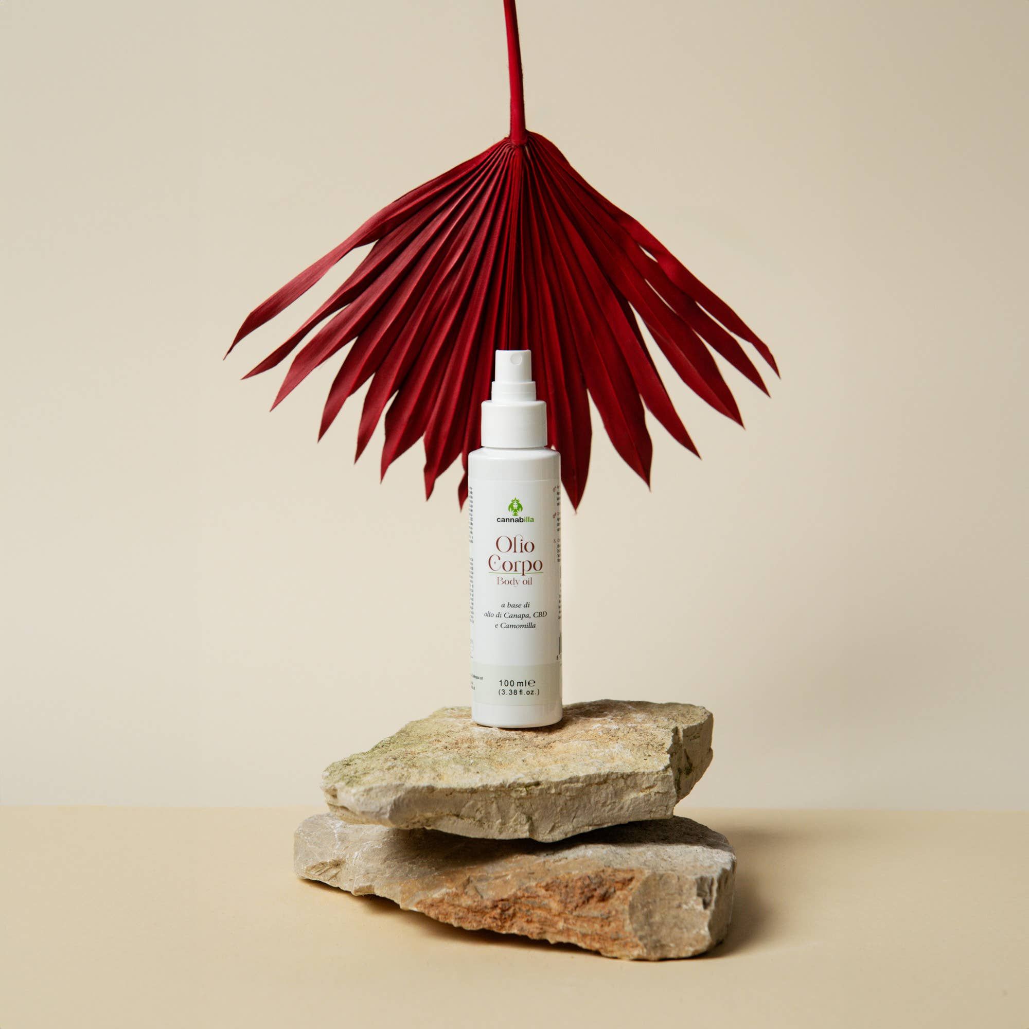 Flacone bianco con erogatore spray su pietre. Scritta: Cannabilla, Olio Corpo Body oil. Foglia rossa a ventaglio sullo sfondo.