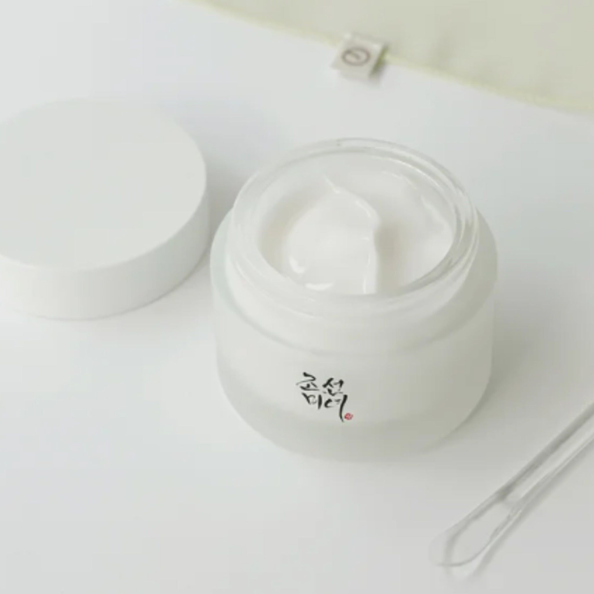 Vasetto di crema aperto, coperchio accanto. Scritta coreana. Spatola trasparente.