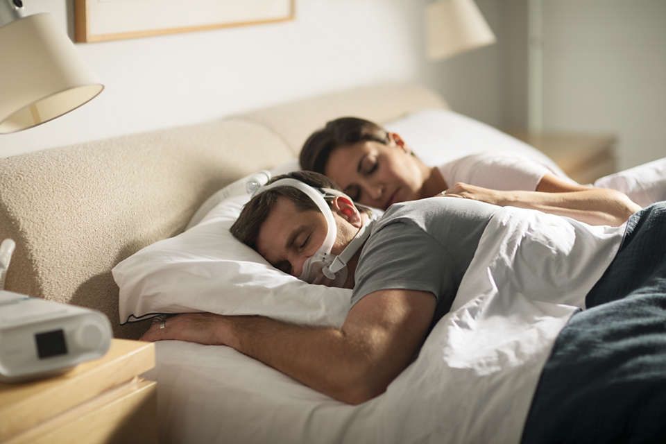 Un uomo dorme a letto con maschera CPAP, una donna dorme accanto. Dispositivo CPAP sul comodino.