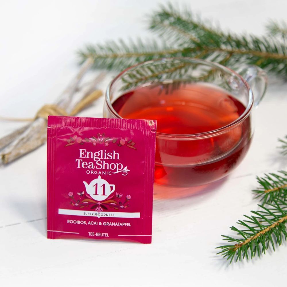 Bustina di tè con "11 Rooibos, Acai & Melograno". Marchio: English Tea Shop. Tè in vetro. Bicchiere con tè rosso. Rametto verde accanto.