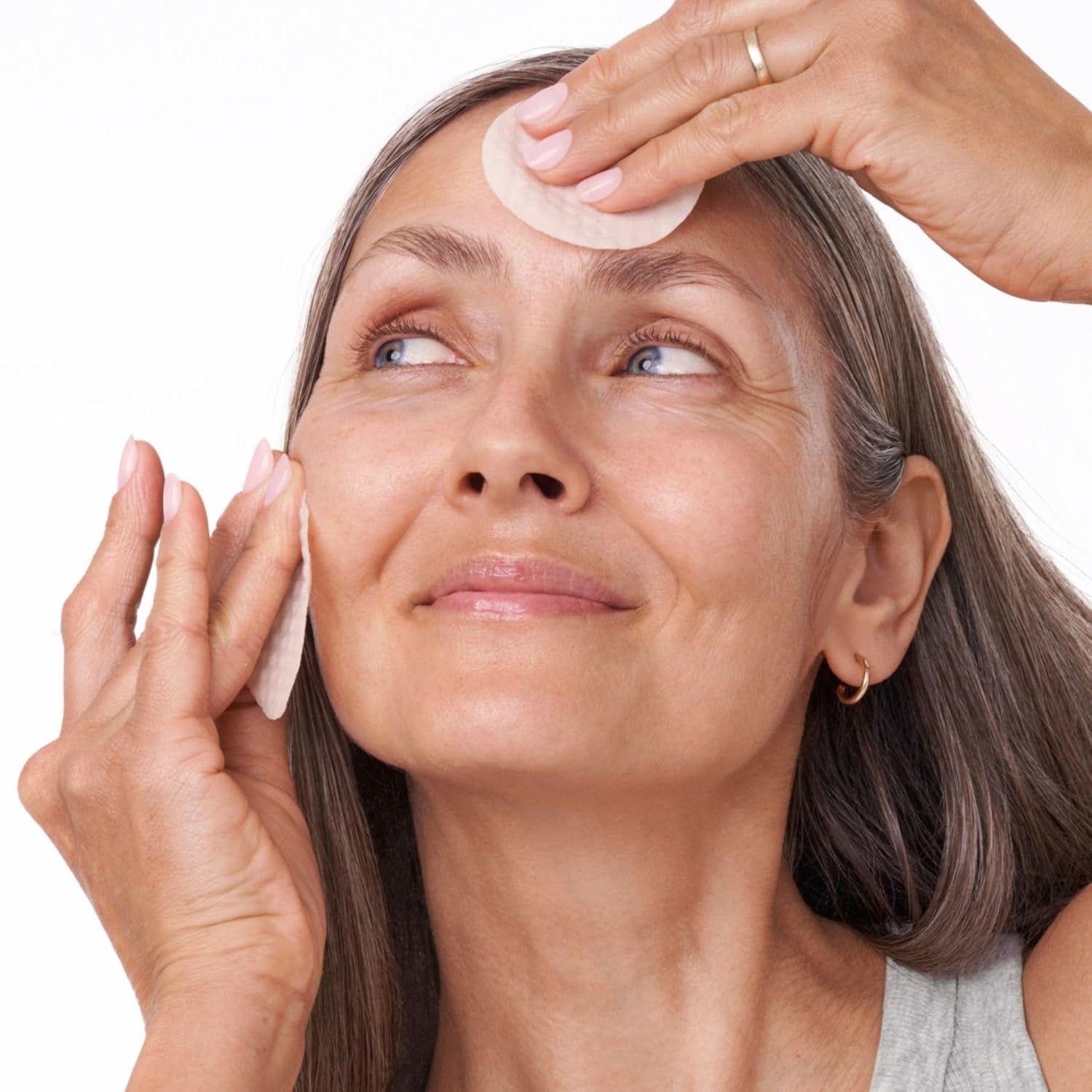 Donna con dischetti per il viso. Mani che tengono i dischetti sulla fronte e sulla guancia. Capelli grigi, top grigio. Guarda in alto.