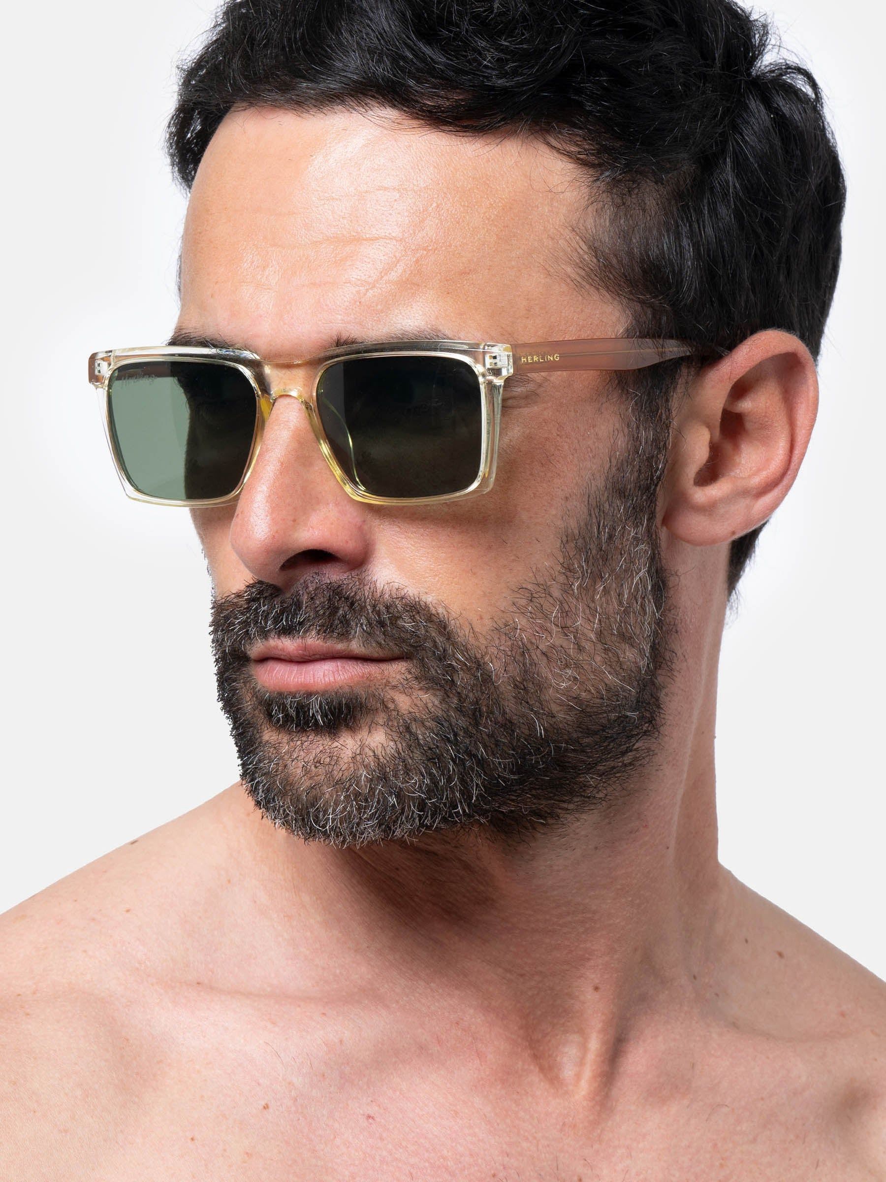 Uomo con barba indossa occhiali da sole con montatura trasparente e lenti verdi. Il marchio 'Herling' è visibile sulla stanghetta.