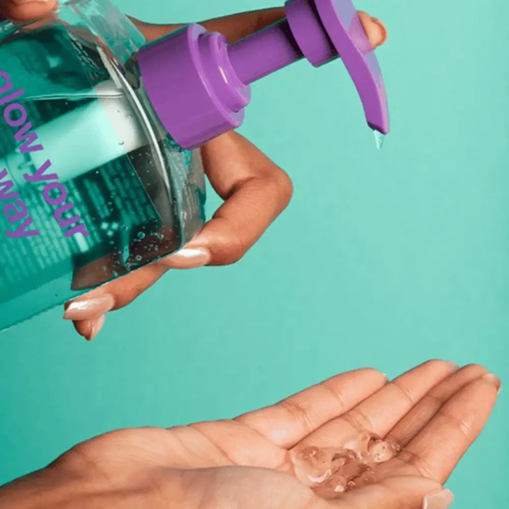 Mano che versa liquido da una bottiglia con pompa viola in un'altra mano. Sulla bottiglia c'è scritto "glow your own way".