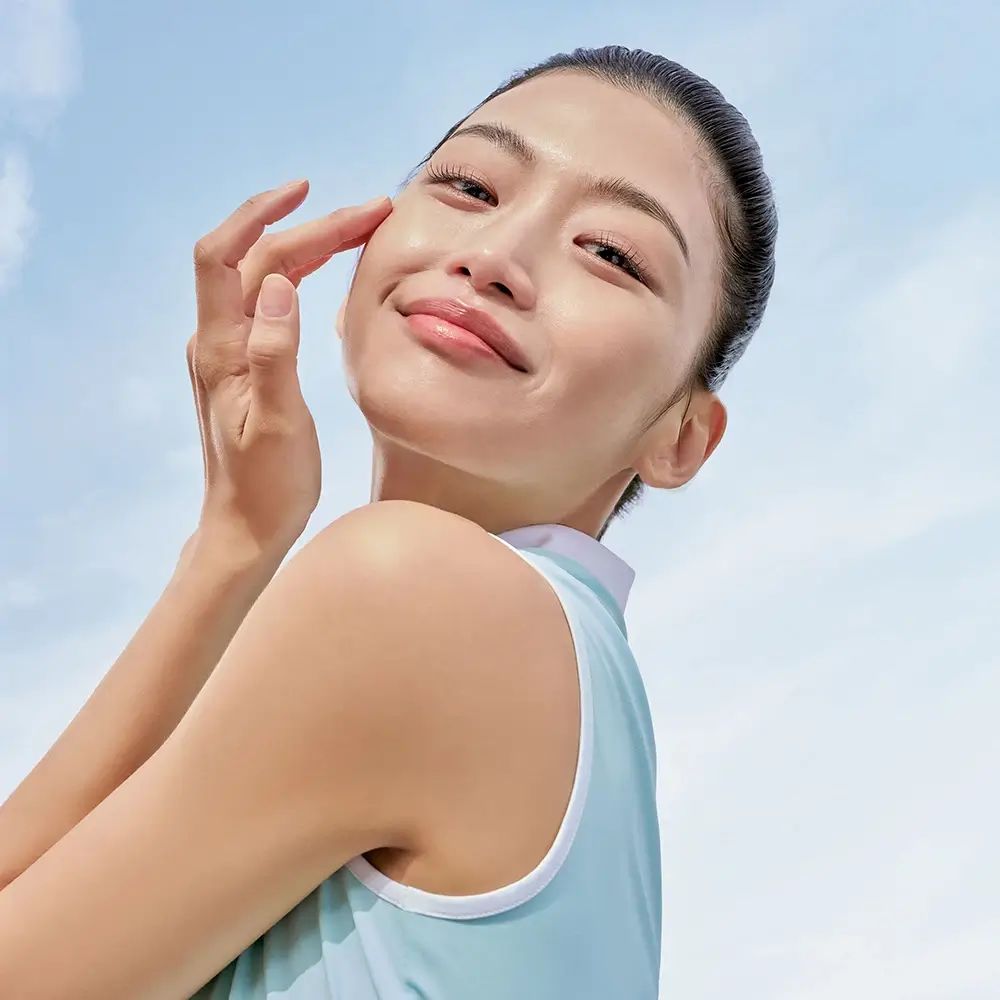 Donna tocca il viso. Cielo azzurro sullo sfondo. Indossa un top azzurro chiaro.