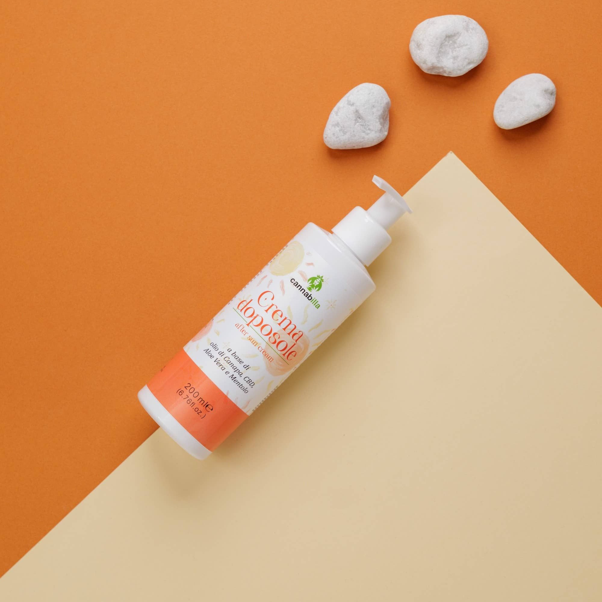 Flacone bianco con dosatore su una superficie inclinata. Scritta: Crema doposole, Cannabilla. Sfondo: arancione e beige. 3 pietre.
