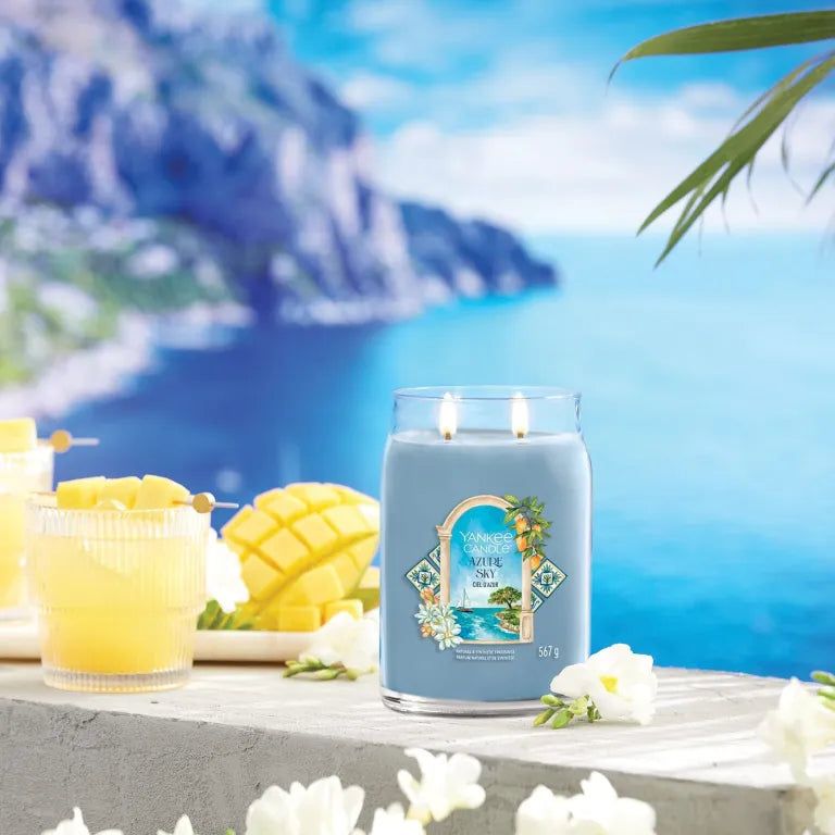 Yankee Candle Azur Sky Signature Grand Pot blu con due stoppini. Scritta sull'etichetta. Sfondo: mare e cocktail.