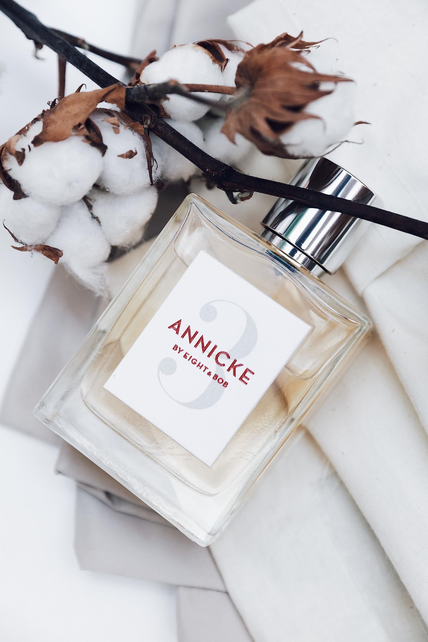 Flacone di profumo con tappo argentato, circondato da rami di cotone e tessuto. Etichetta "ANNICKE BY EIGHT & BOB" e numero "3".