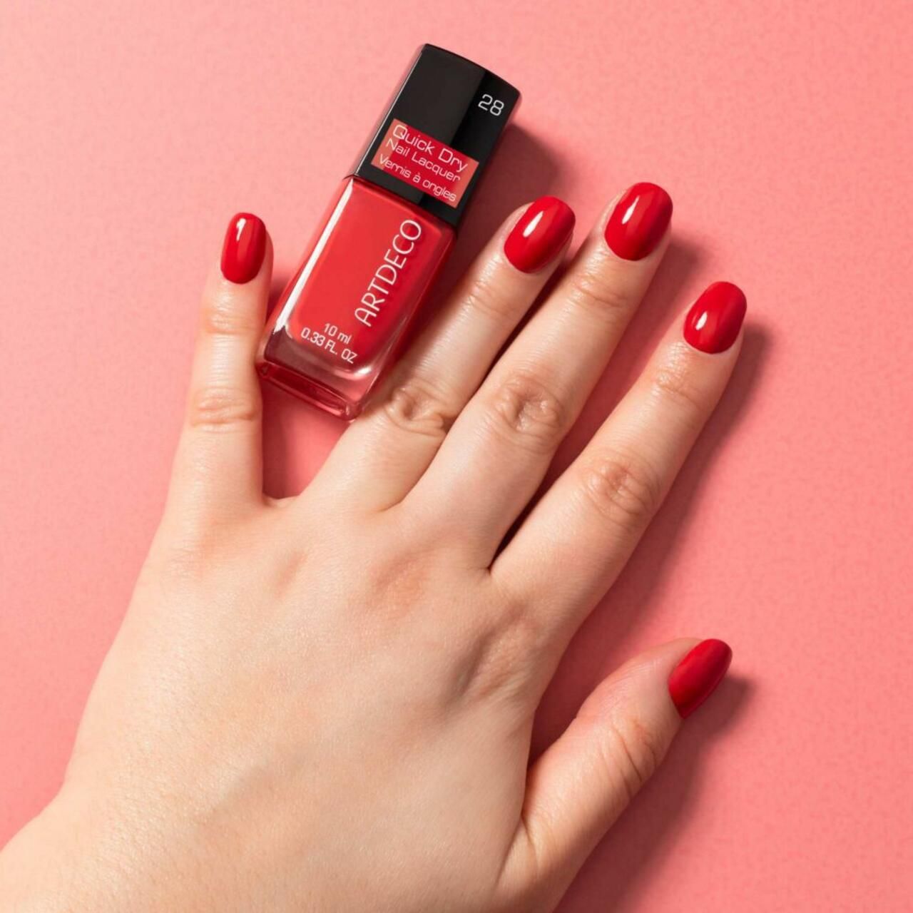 Mano con unghie rosse smaltate accanto a un flacone di smalto per unghie Artdeco Quick Dry. Scritta: Quick Dry Nail Lacquer, Vernis à ongles.