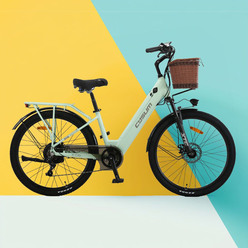 Bicicletta elettrica verde chiaro con cestino, sella nera e pneumatici. Sfondo giallo e blu.