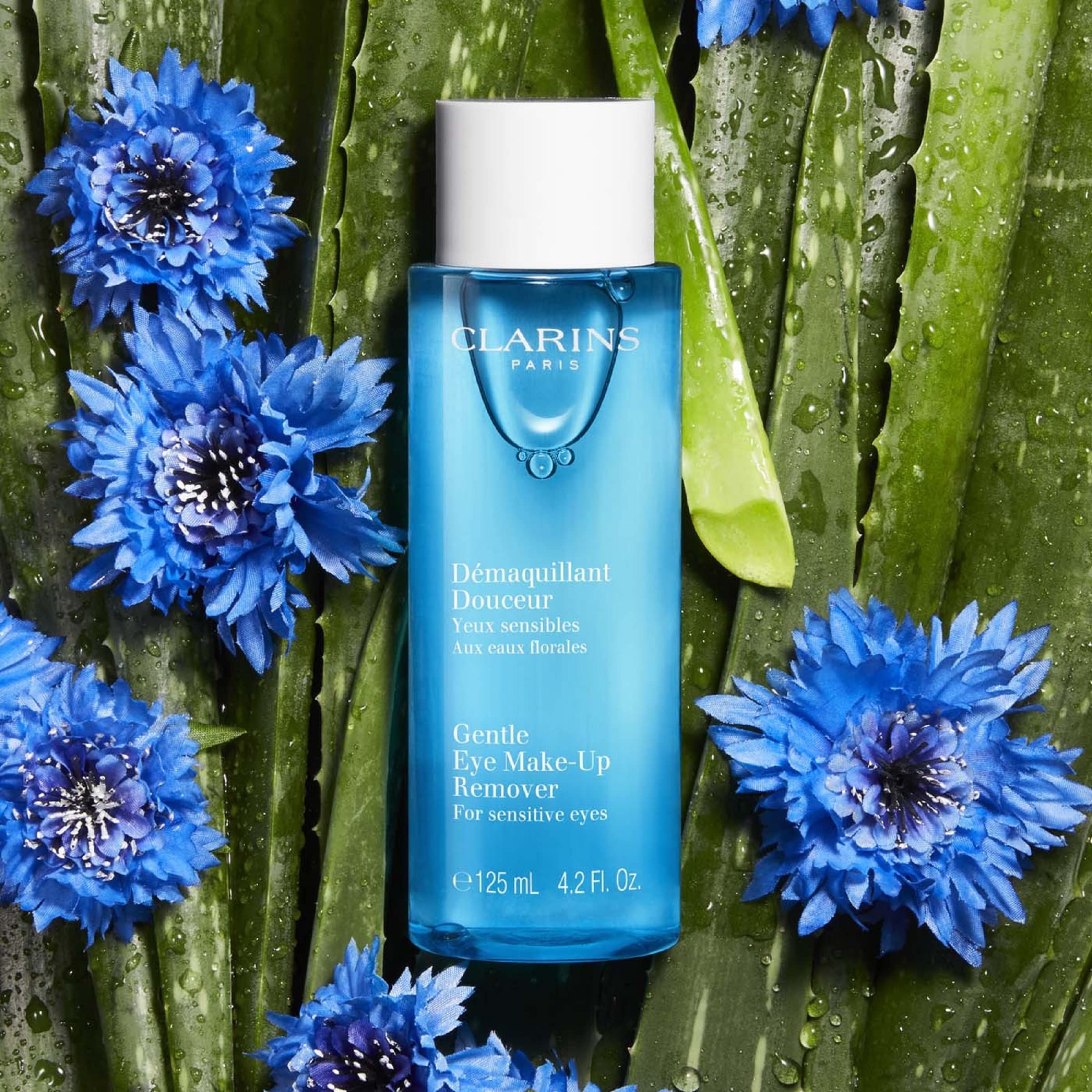 Flacone blu di struccante occhi. Testo: Clarins, Démaquillant Douceur, Gentle Eye Make-up Remover. Circondato da fiori blu e foglie.