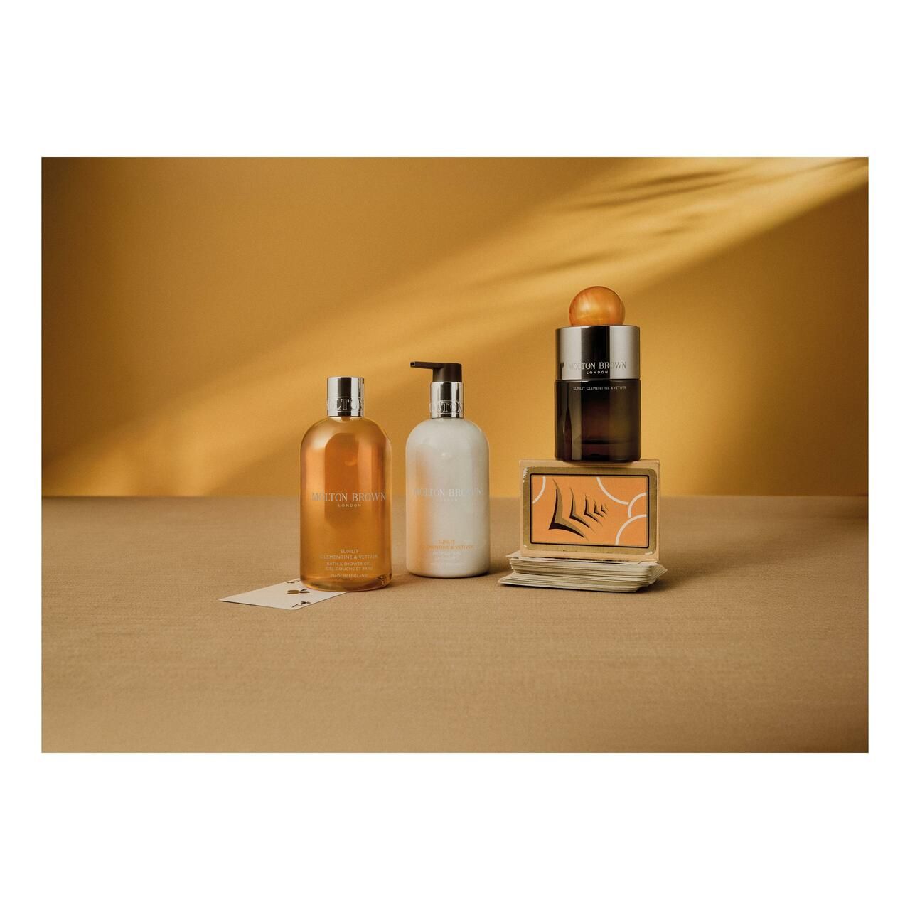 Disposizione con flaconi e una scatola. Prodotti: Molton Brown, Sunlit Clementine & Vetiver. Sfondo: giallo.