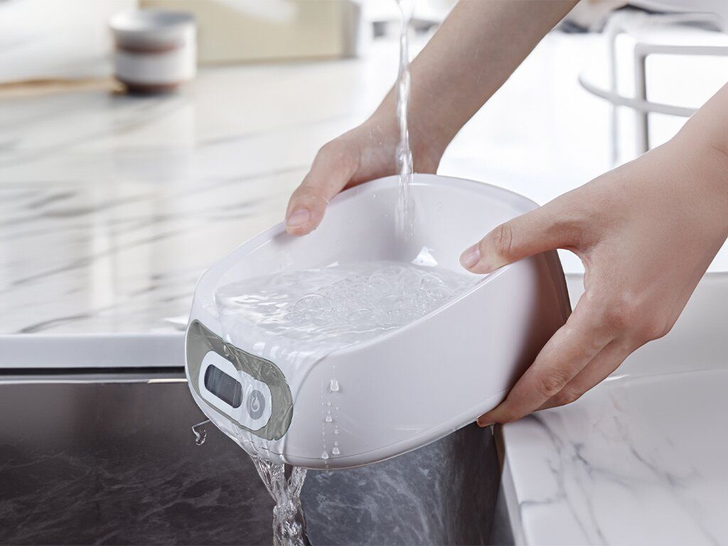 Ciotola bianca con bilancia integrata, riempita d'acqua. La ciotola è tenuta in mano sopra un lavandino.