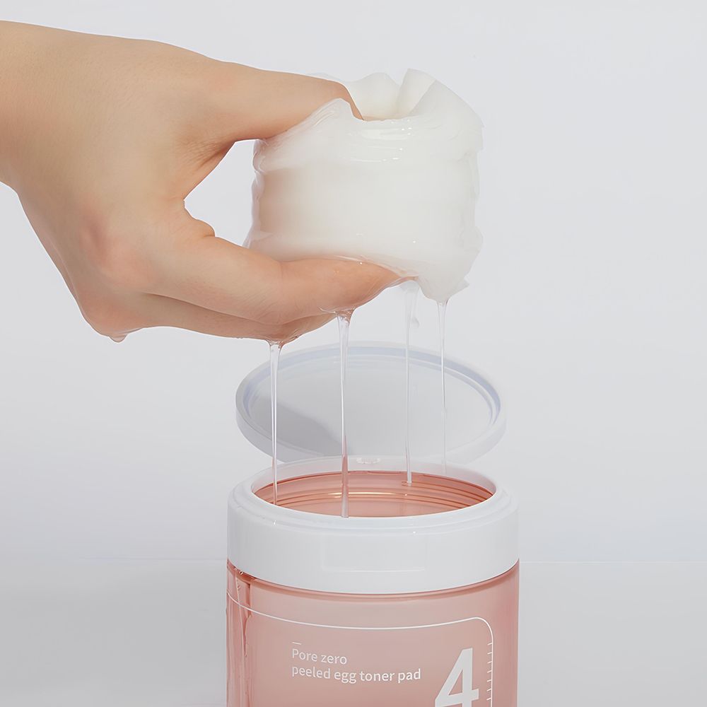 Mano che tiene un pad imbevuto sopra un barattolo aperto. Liquido che gocciola. Barattolo rosa, coperchio bianco. Pad bianchi.