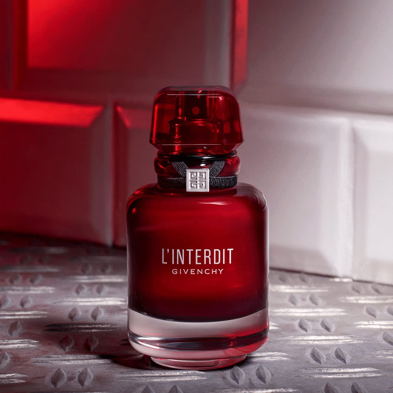 Flacone di profumo rosso con nastro nero e tappo argentato. Scritta: L'Interdit Givenchy. Sfondo: metallo e piastrelle.