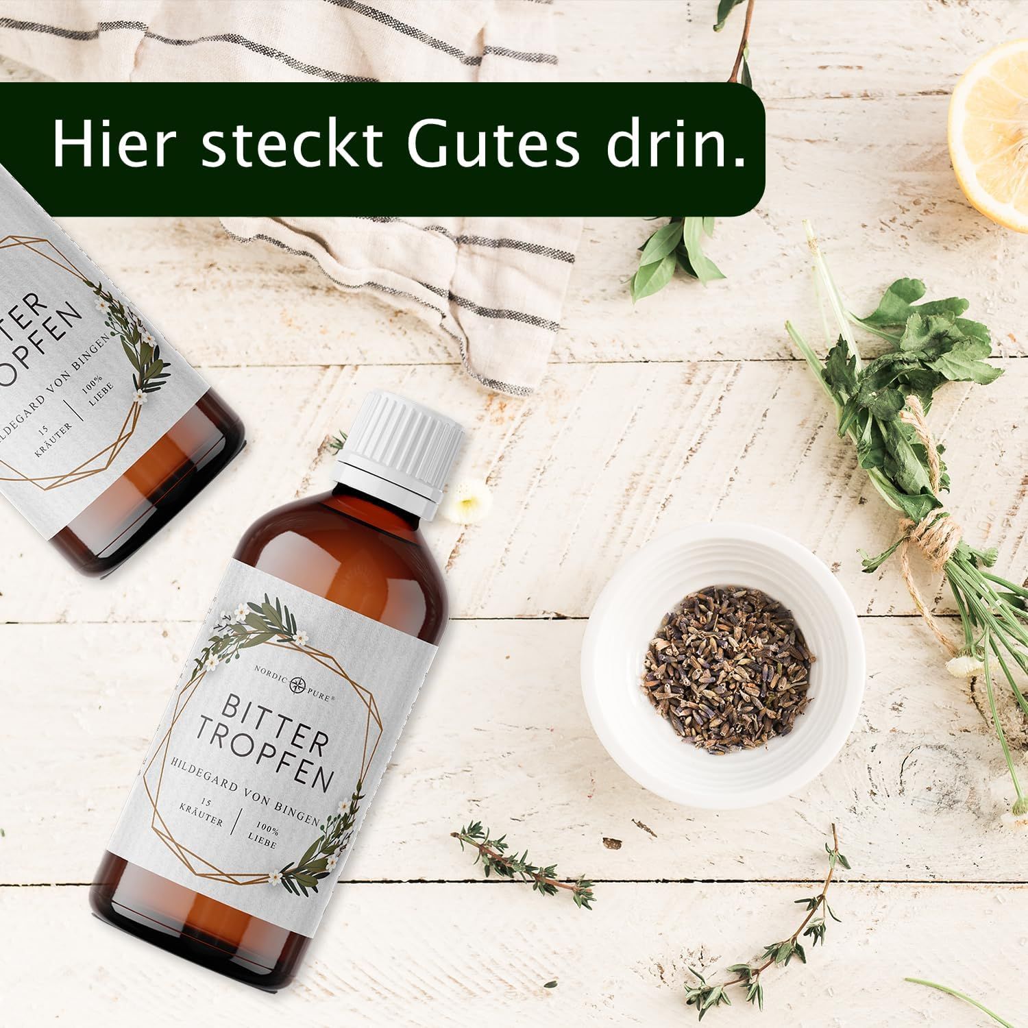 Due bottiglie marroni con tappo bianco. Etichetta con nome del prodotto. Su legno. Erbe e limone. Testo: C'è del buono qui dentro.