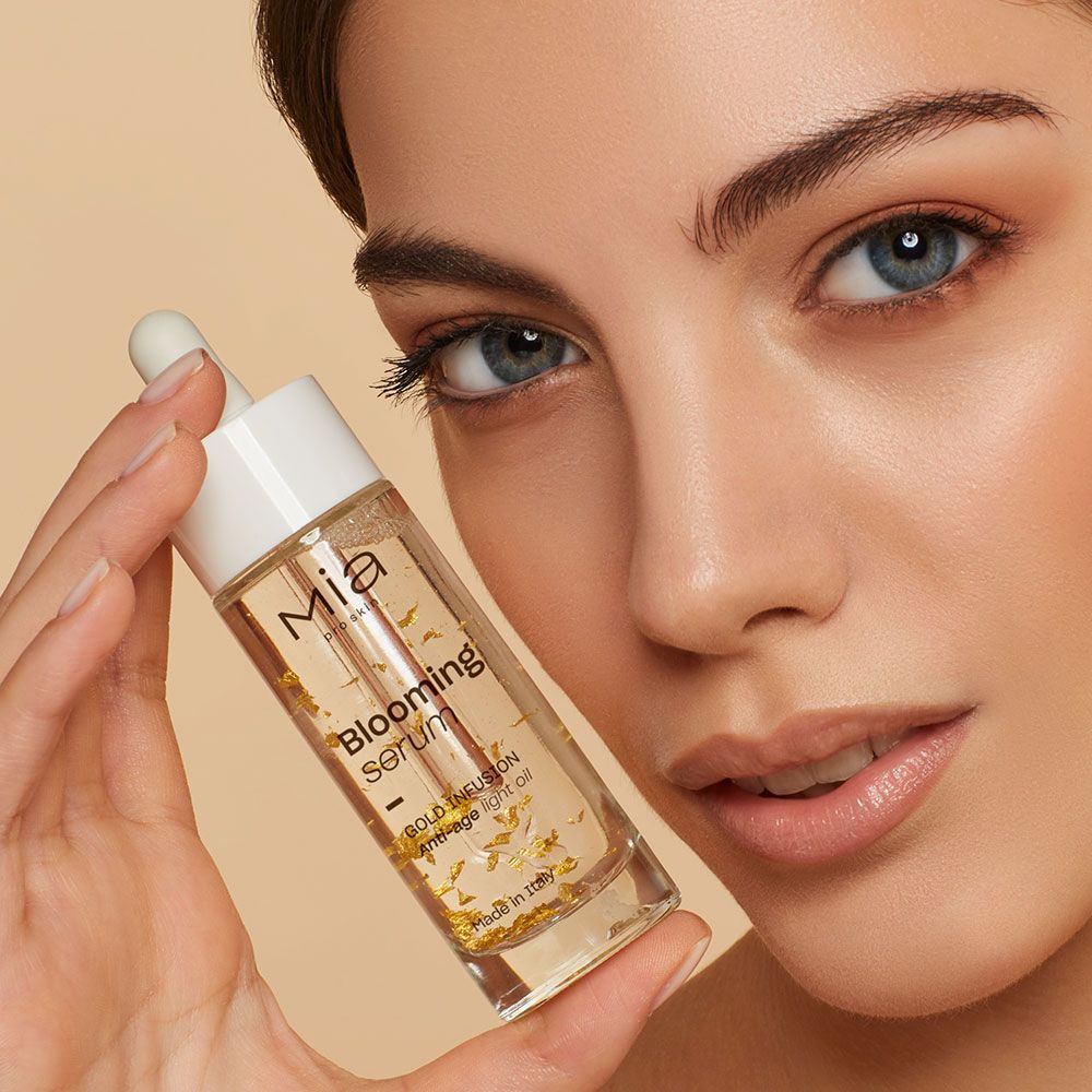 Donna tiene in mano flacone di siero. Siero trasparente con particelle d'oro. Scritte: Mia, Blooming Serum, Gold Infusion, Anti-Age Light Oil.