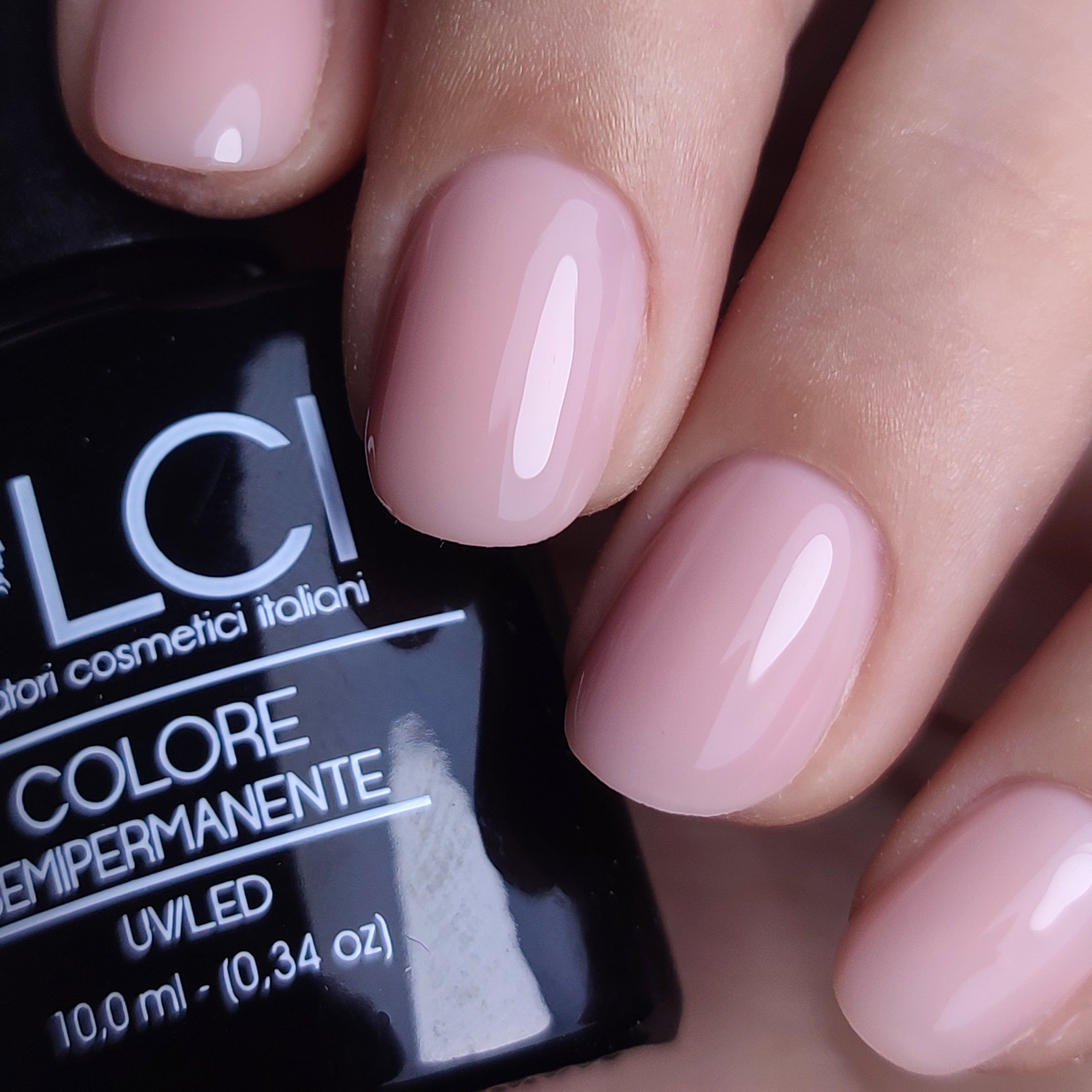 Primo piano di unghie smaltate. Colore rosa chiaro. Flacone con nome del prodotto e logo sullo sfondo.