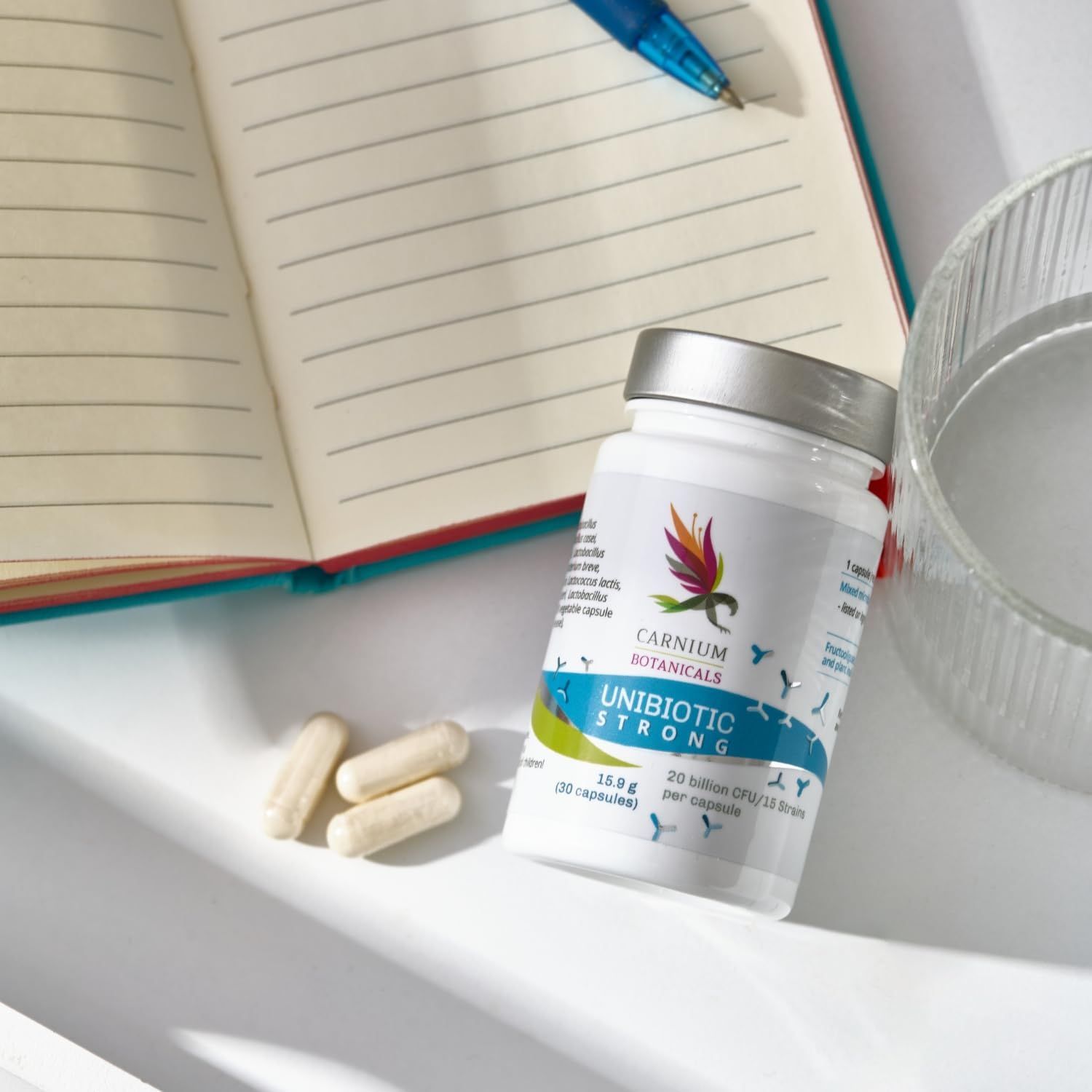 Flacone con capsule vicino a taccuino e bicchiere. Scritta: Unibiotic STRONG, 20 miliardi di CFU/15 ceppi. Marchio: CARNIUM BOTANICALS.