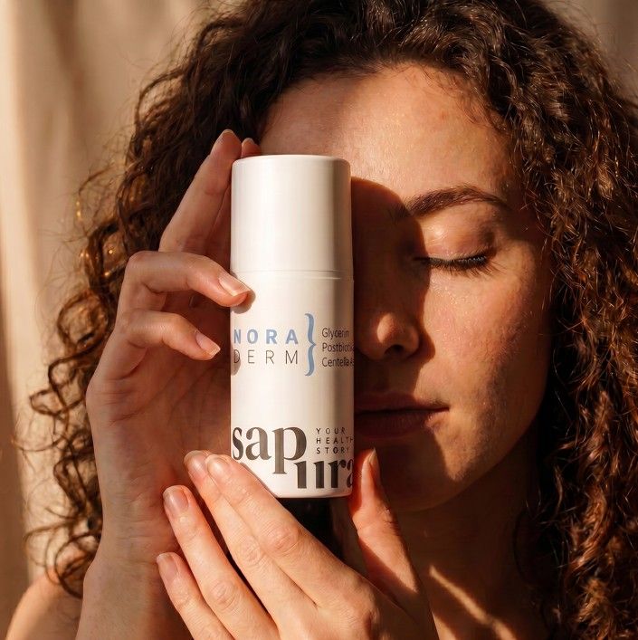 Flacone dispenser bianco con i loghi 'Nora Derm' e 'SAPURA'. Una donna tiene il flacone davanti al viso.