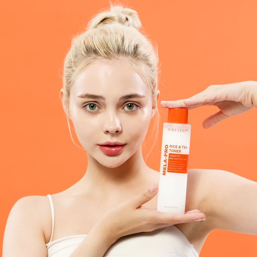 Una donna tiene in mano una bottiglia bianca con tappo arancione. La bottiglia reca la scritta "NINE LESS Mela-Pro Rice & TXA Toner".