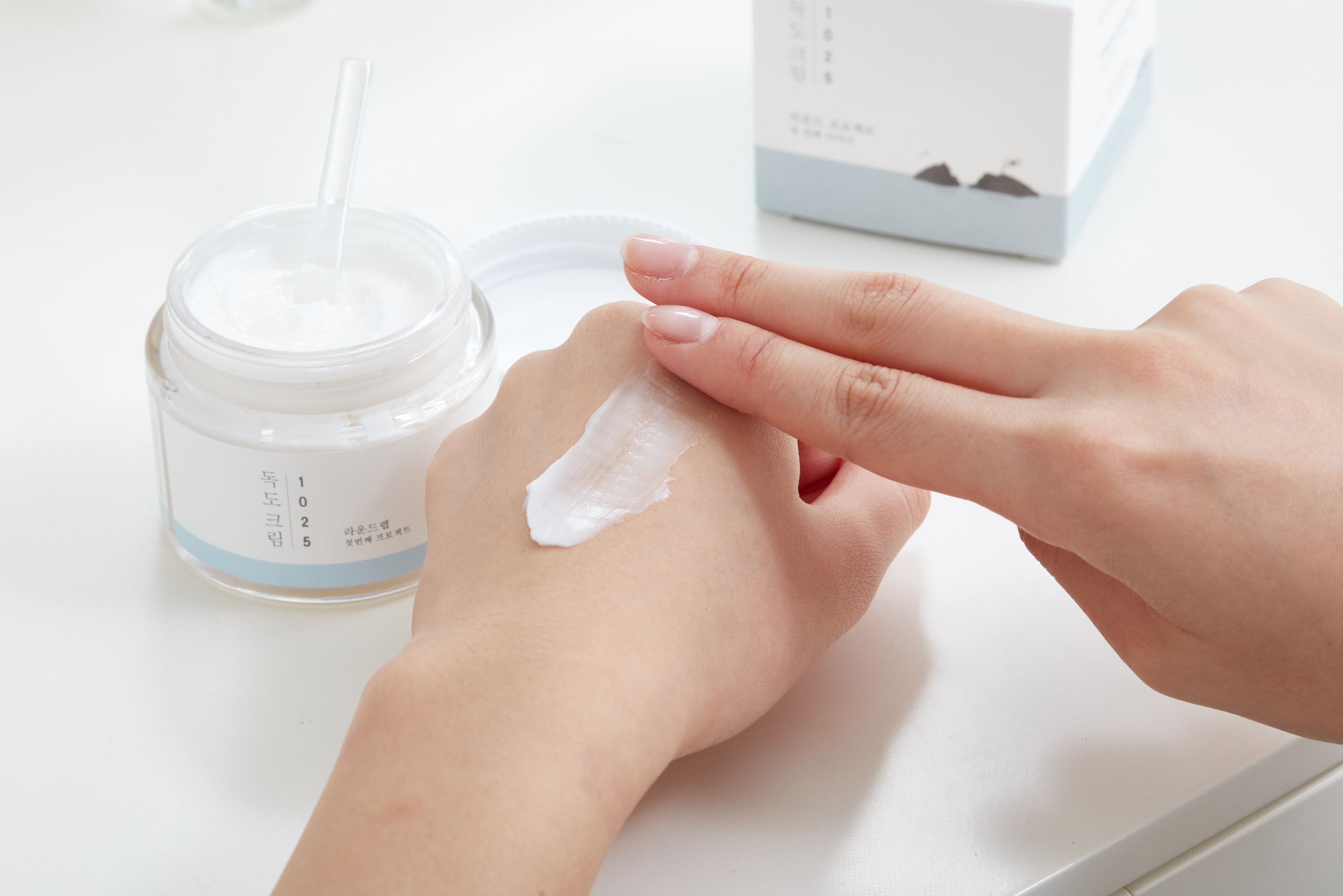 Mani che applicano crema. Vasetto di crema aperto e spatola accanto. Crema bianca sulla pelle.