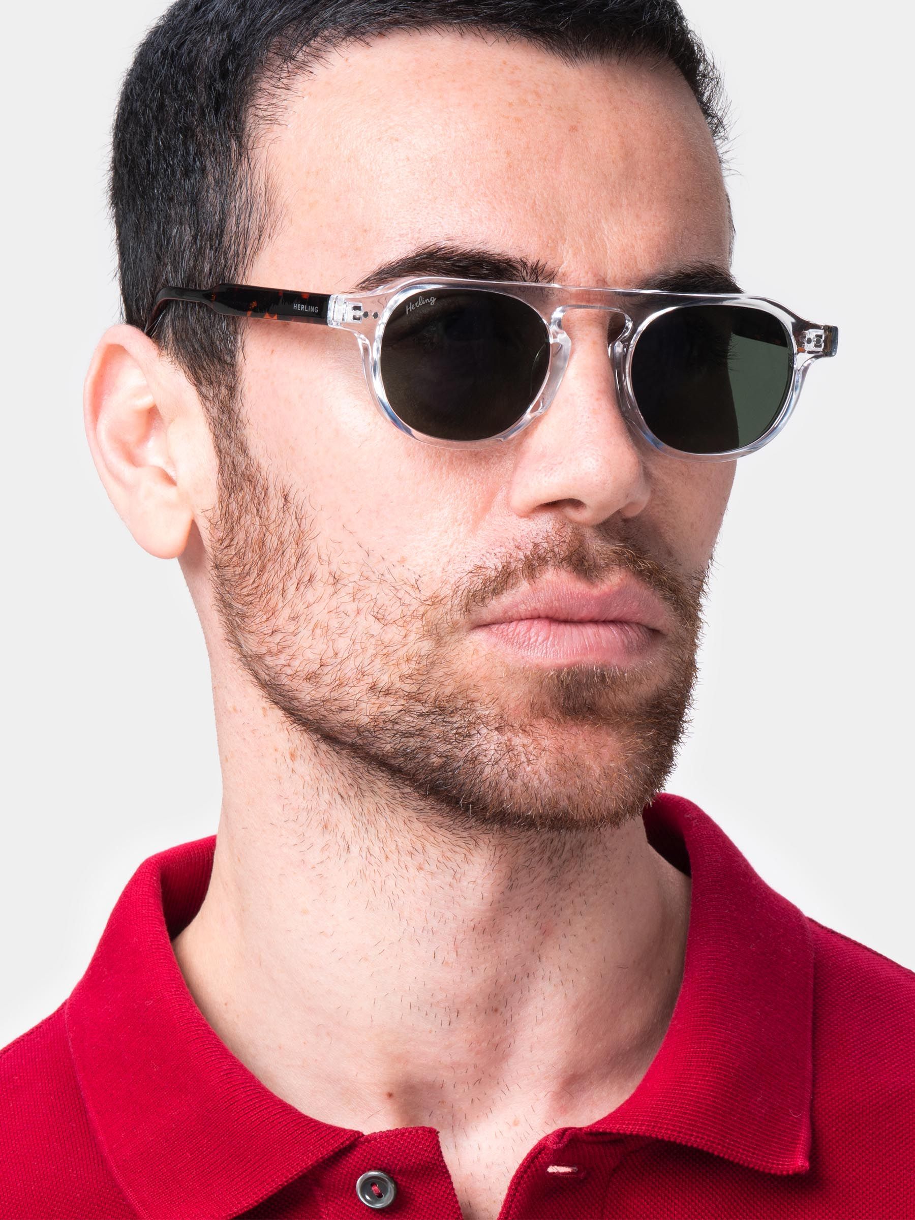 Primo piano di uomo con occhiali da sole. Montatura trasparente, lenti scure. Logo Herling. Polo rosso.