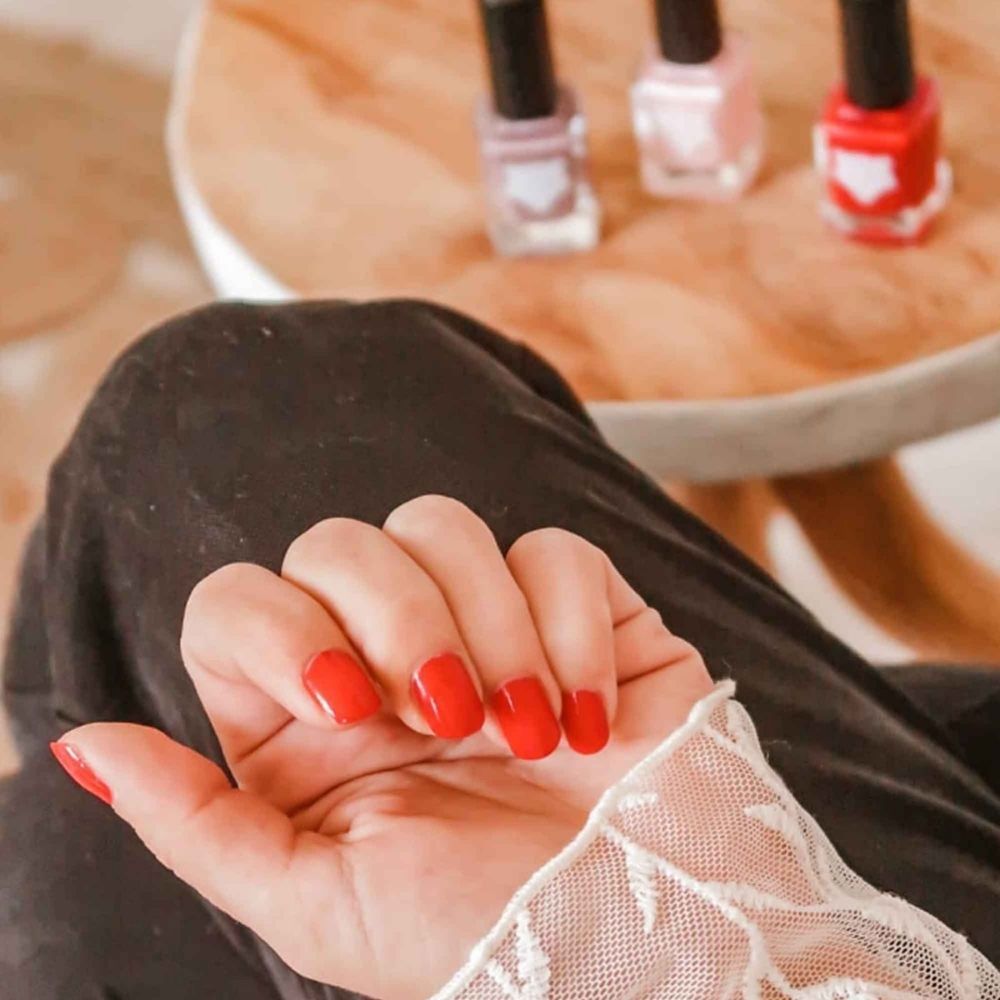 Mano con unghie rosse. Sullo sfondo: tre flaconi di smalto.