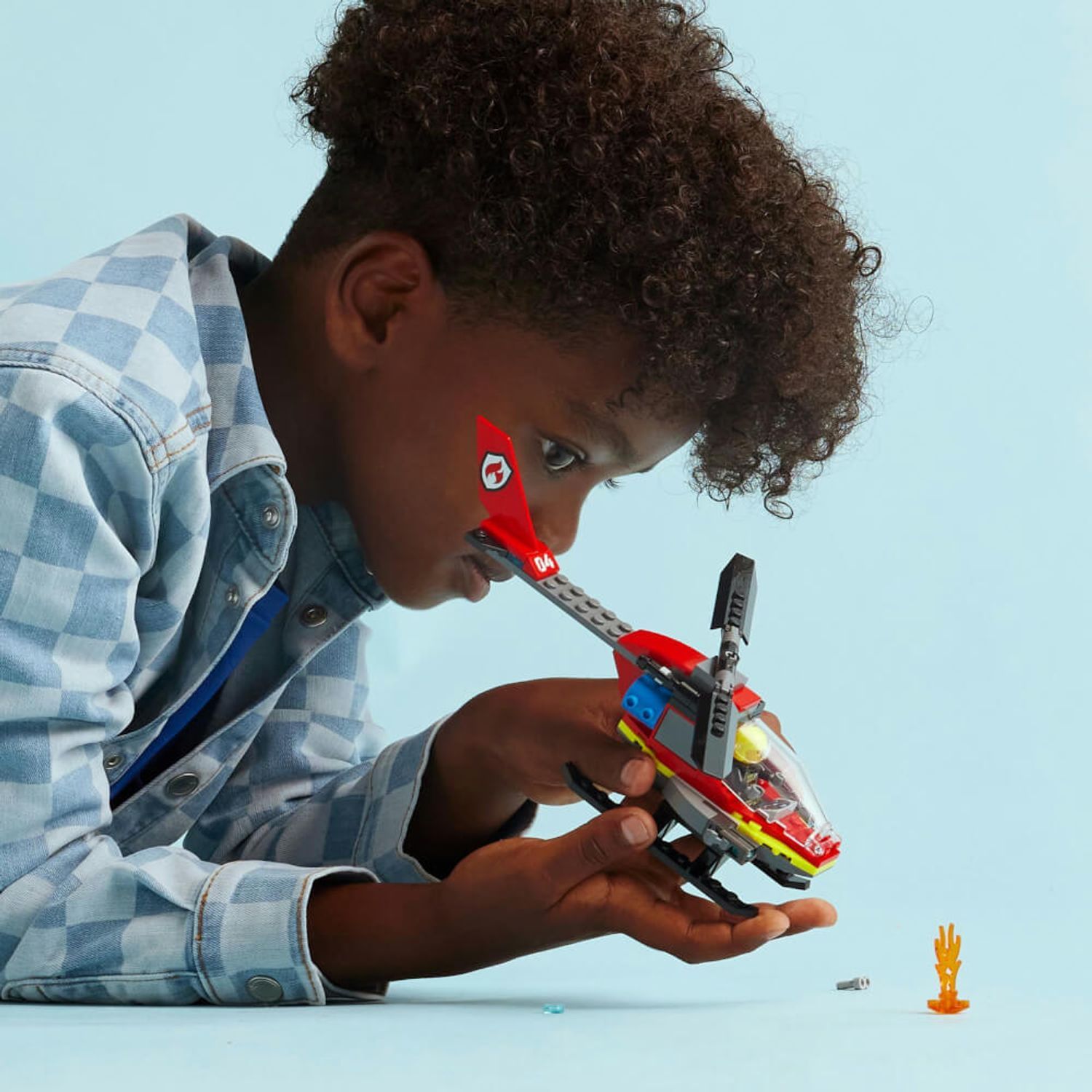 Un bambino tiene in mano un elicottero dei pompieri giocattolo rosso e giallo. Accanto a lui ci sono un elemento fiamma e un piccolo pezzo grigio.