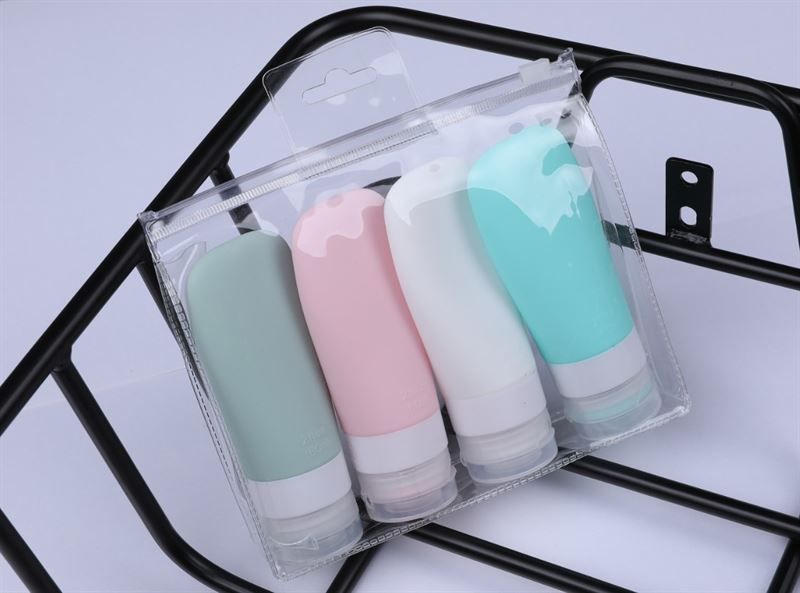 Quattro flaconi da viaggio colorati in una borsa trasparente. Colori: verde, rosa, bianco e turchese. Su un supporto nero.