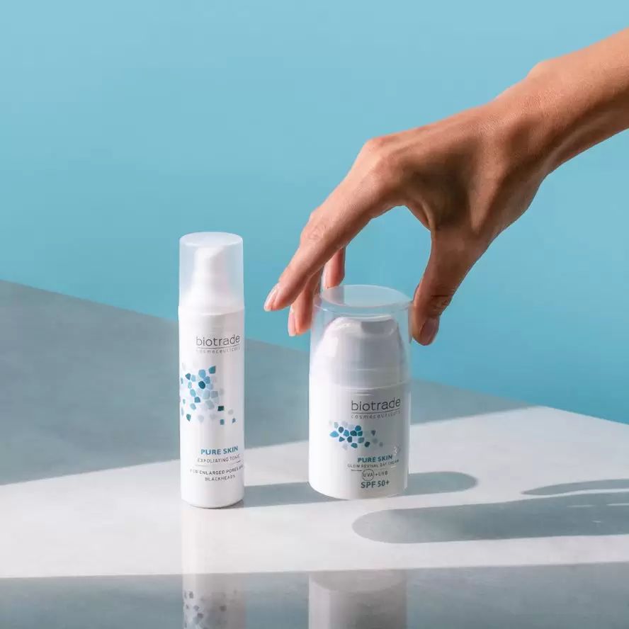 Due flaconi di crema bianchi. Scritta: Biotrade, Pure Skin, SPF 50+. Una mano si protende verso un flacone.