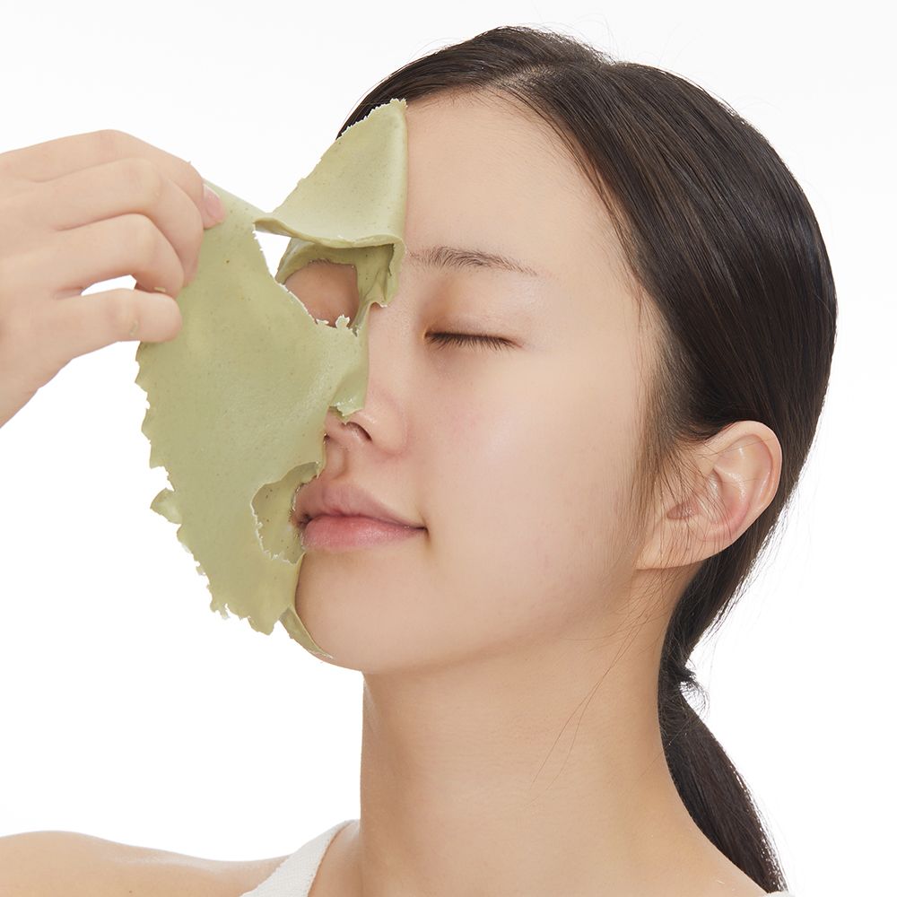 Viso di donna con maschera verde. Una mano rimuove la maschera. Sfondo bianco.