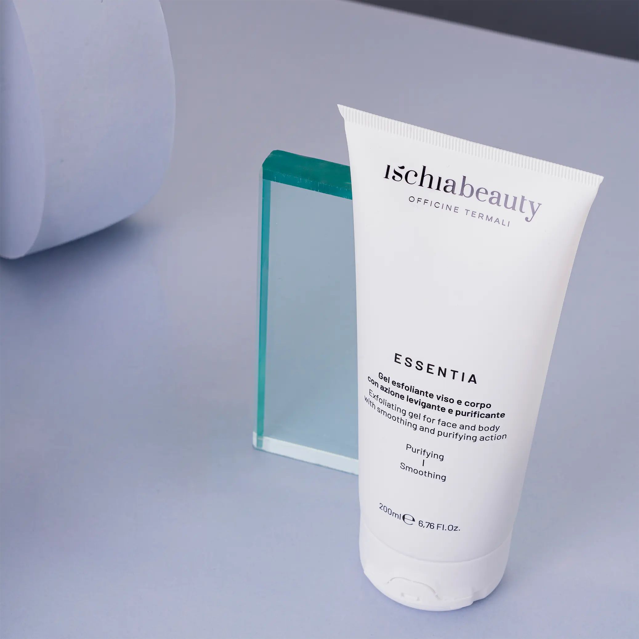Tubo bianco con testo del prodotto. Testo: Ischiabeauty, Essentia, Gel esfoliante viso e corpo. Su sfondo blu, accanto a oggetto in vetro.
