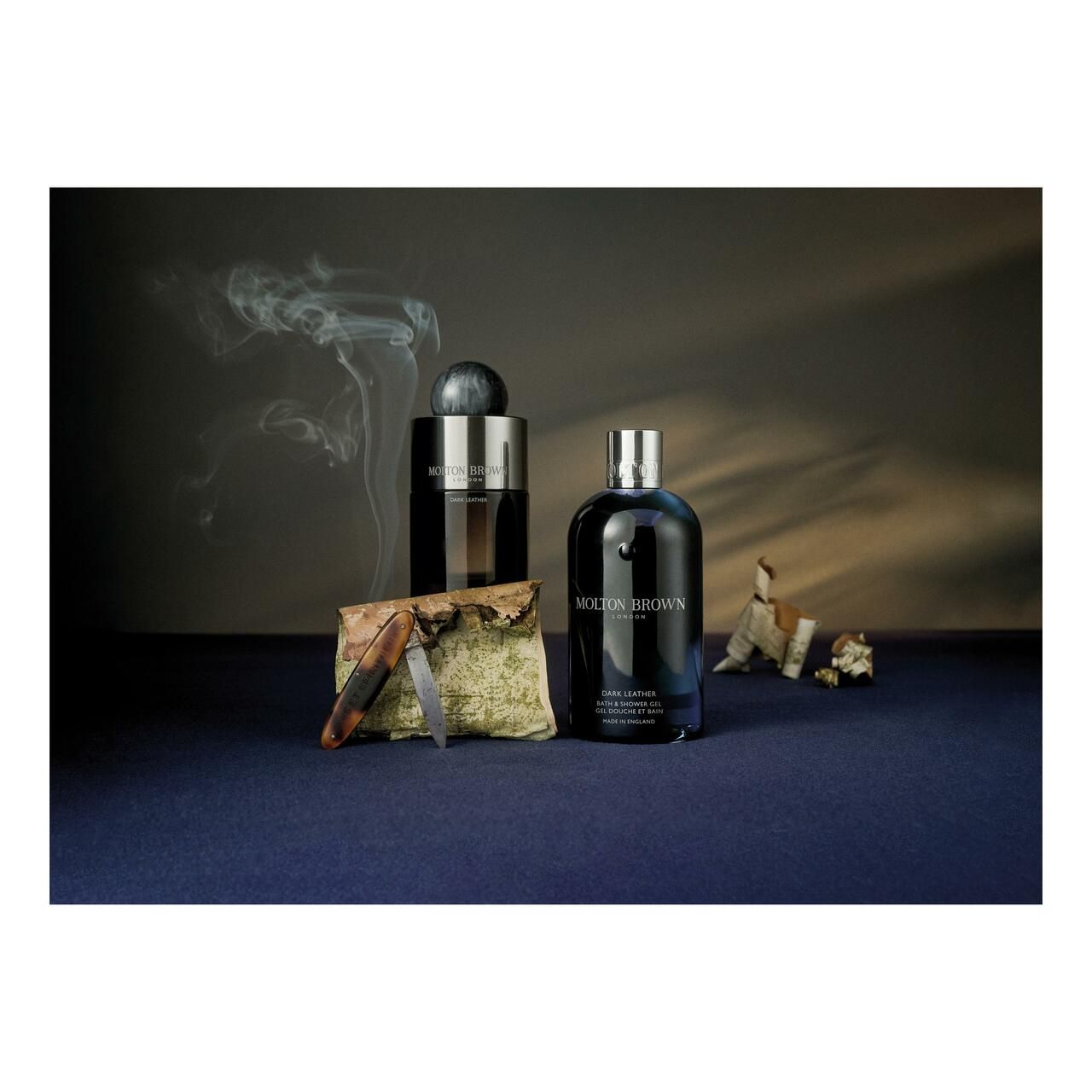 Flacone blu scuro, accanto a un altro flacone e accessori su sfondo blu. Fumo sullo sfondo.