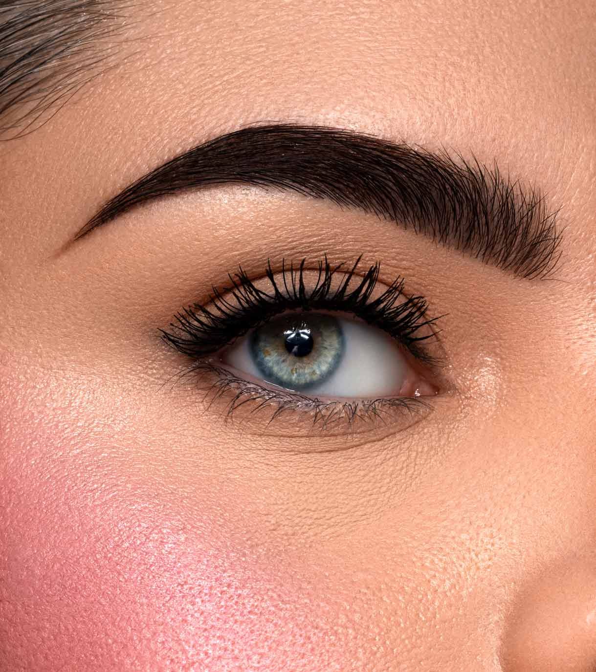 Primo piano di un occhio umano con trucco scuro e arcuato per le sopracciglia e ciglia folte. La pelle è accentuata da un fard rosa.