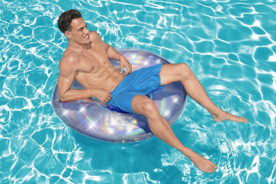 Uomo seduto in un anello da nuoto iridescente in piscina. Argento con riflessi arcobaleno. Pantaloncini blu.