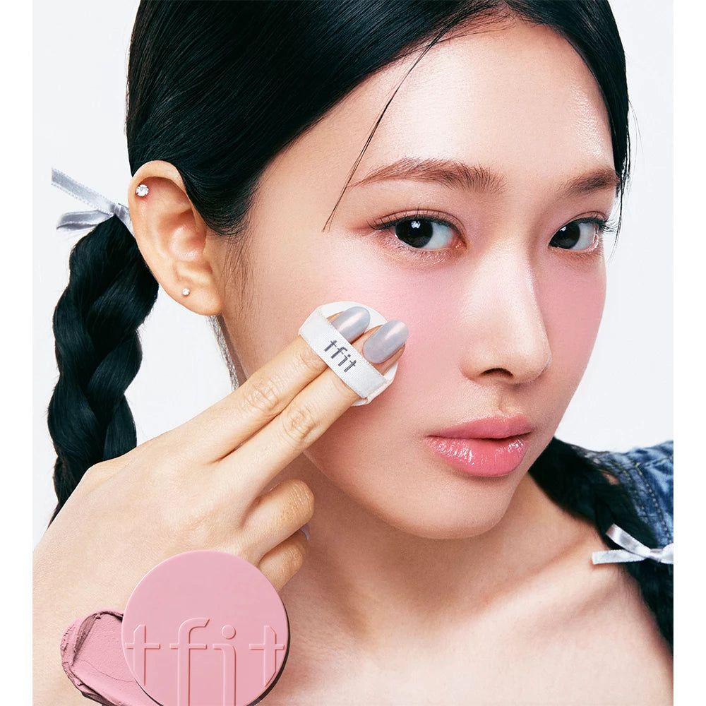 Persona con trecce applica TFIT Fluffy Velvet Cushion Blush con applicatore. Fard rosa sulla guancia visibile. Nome del prodotto sull'applicatore.