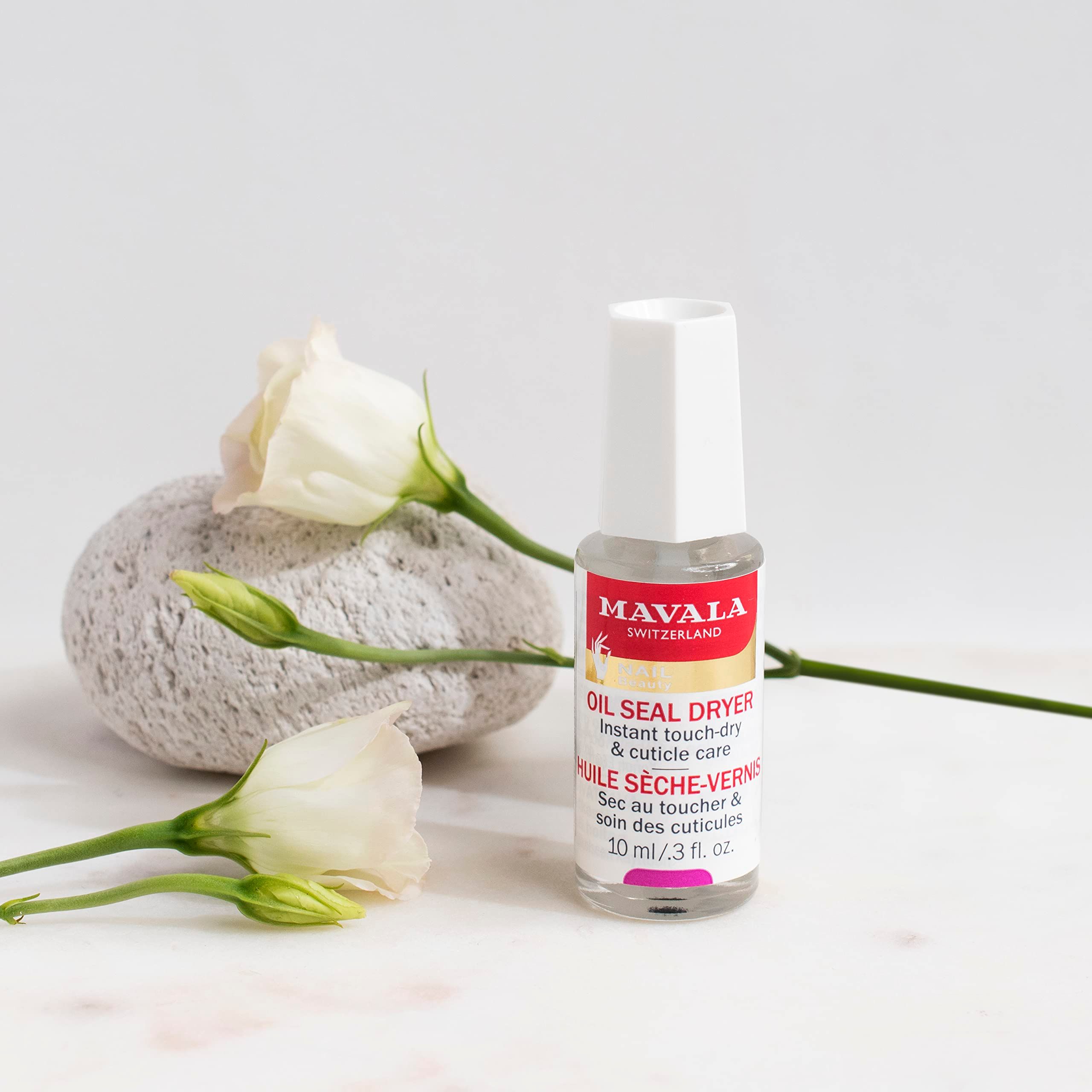 Piccolo flacone con etichetta del prodotto. Marchio: Mavala. Su sfondo bianco con fiori e pietra.