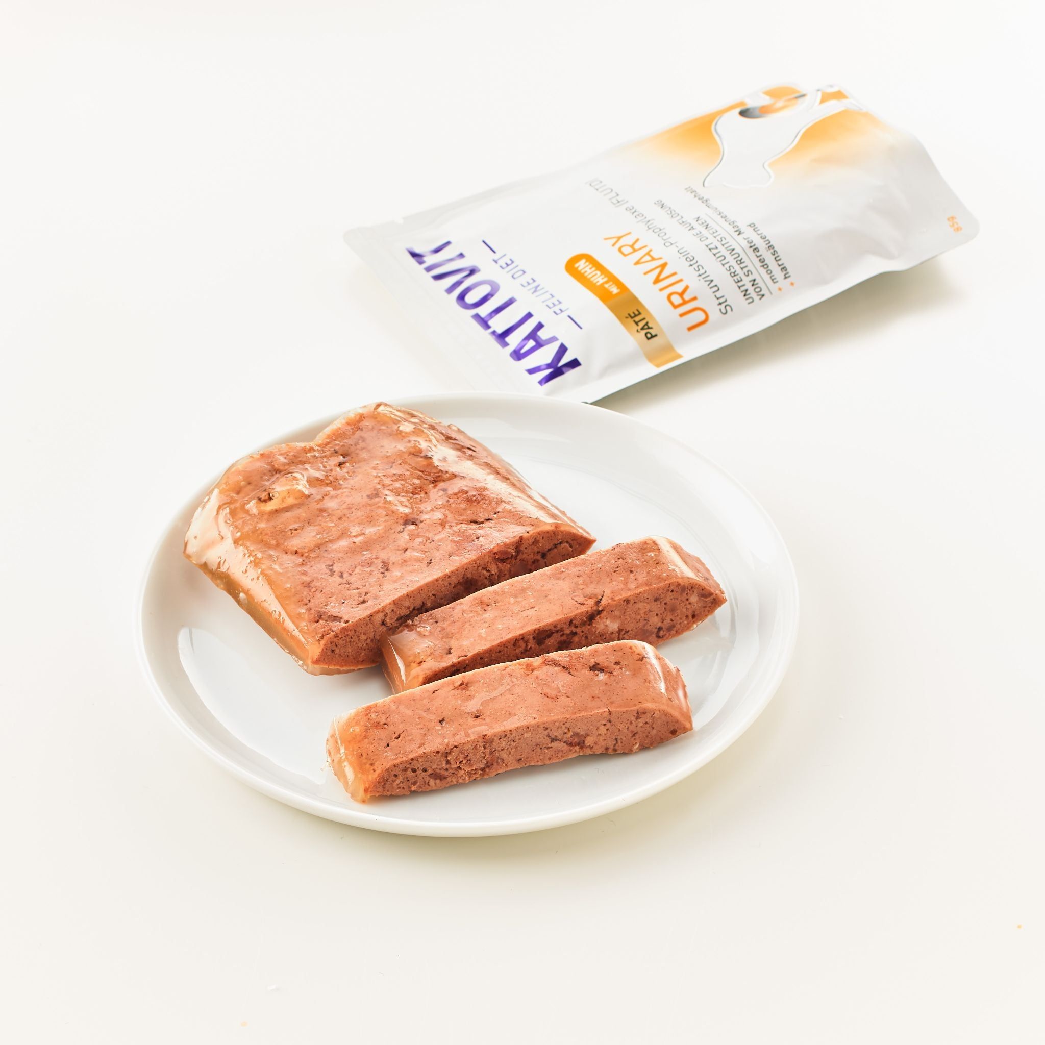 KATTOVIT Urinary Paté Huhn su un piatto. Cibo modellato, tagliato a fette. Confezione sullo sfondo.