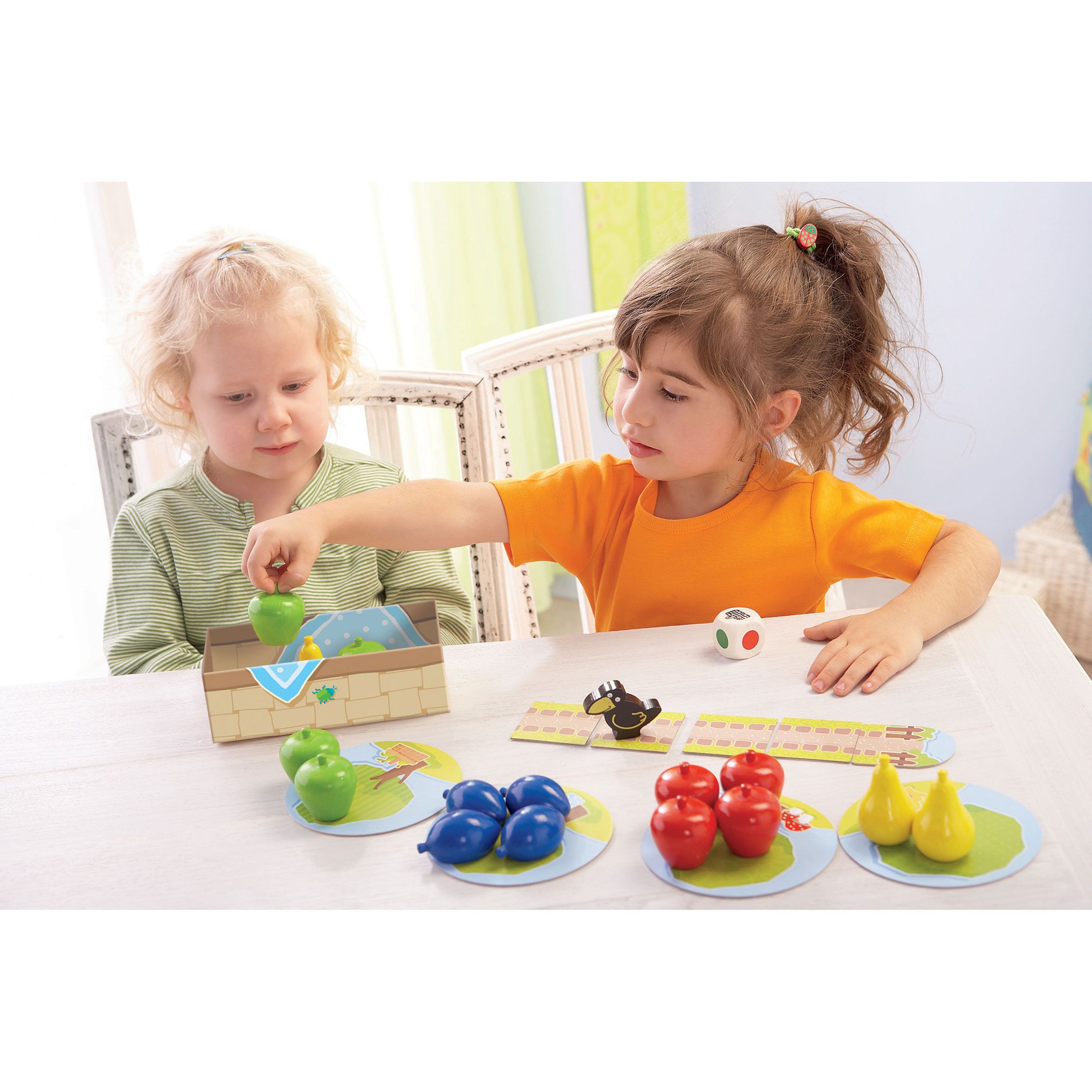 Due bambini giocano. Tabellone, frutta, dadi e cesto sul tavolo. Un bambino mette la frutta nel cesto.