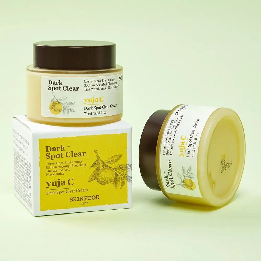 Due vasetti di crema e una scatola. Scritta: Dark Spot Clear, Yuja C, SKINFOOD. Crema gialla in un barattolo con coperchio marrone. Scatola con informazioni sul prodotto.