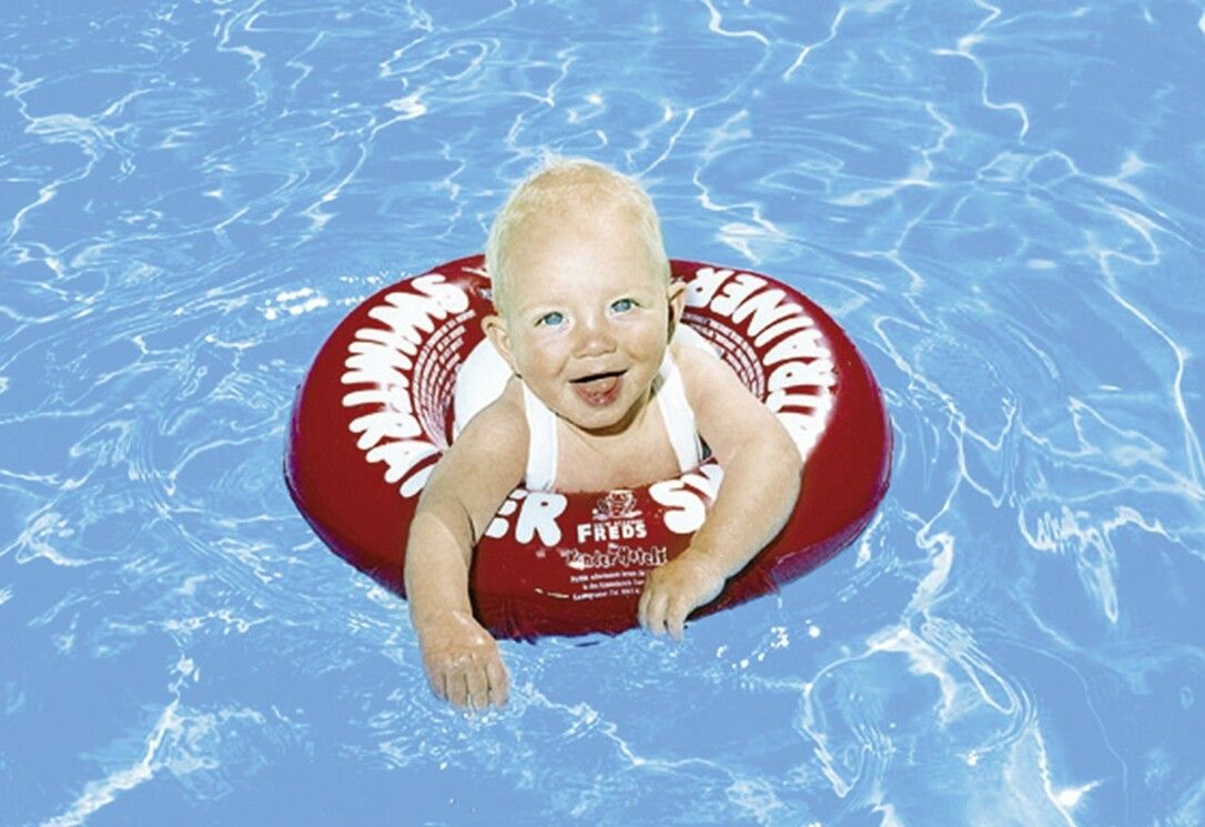 Bambino in Schwimmtrainer rosso in piscina. Marchio Freds Swim Academy. Il bambino sorride.