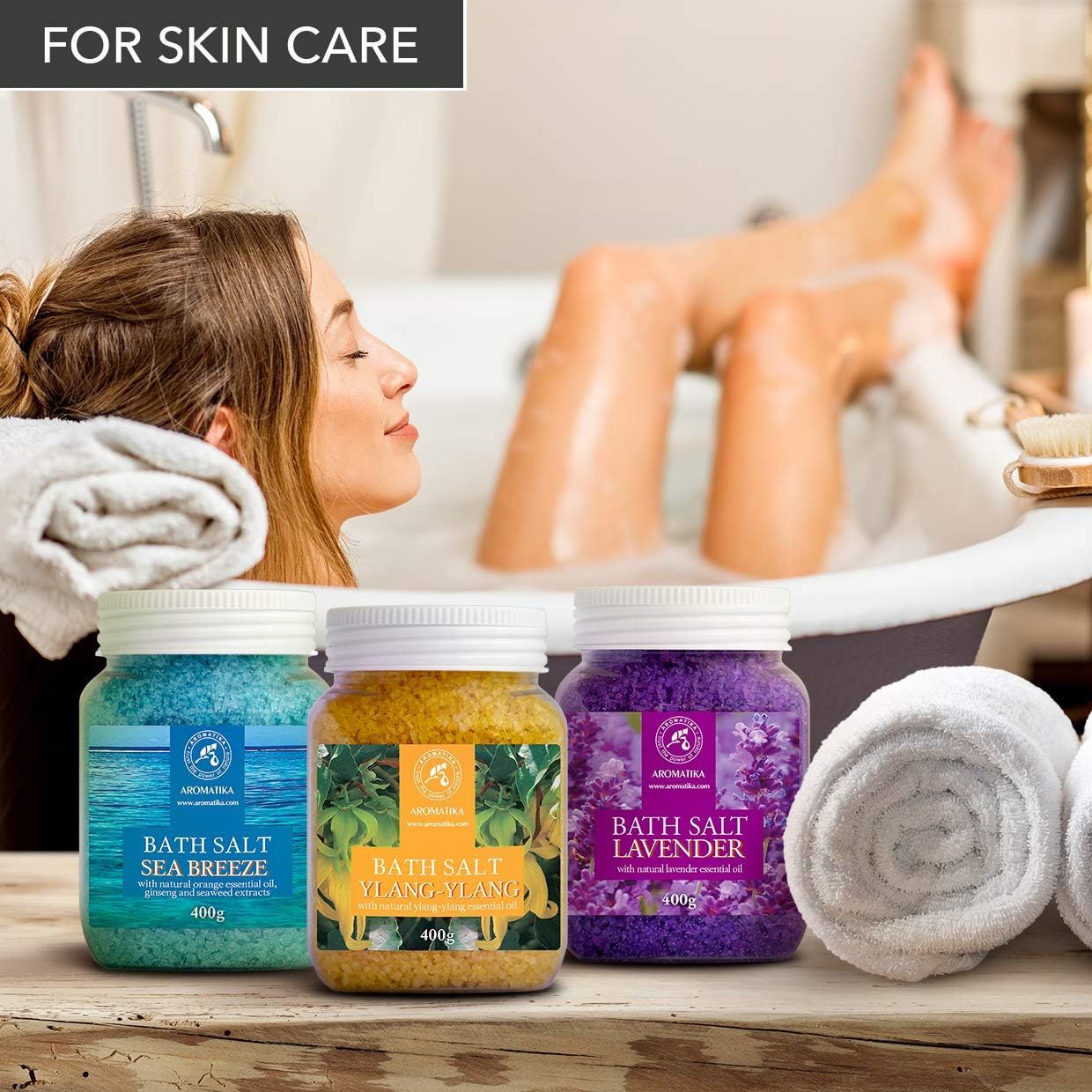 Tre barattoli di sali da bagno accanto a una donna nella vasca. Blu, giallo e viola. Scritte: Sea Breeze, Ylang-Ylang, Lavanda.