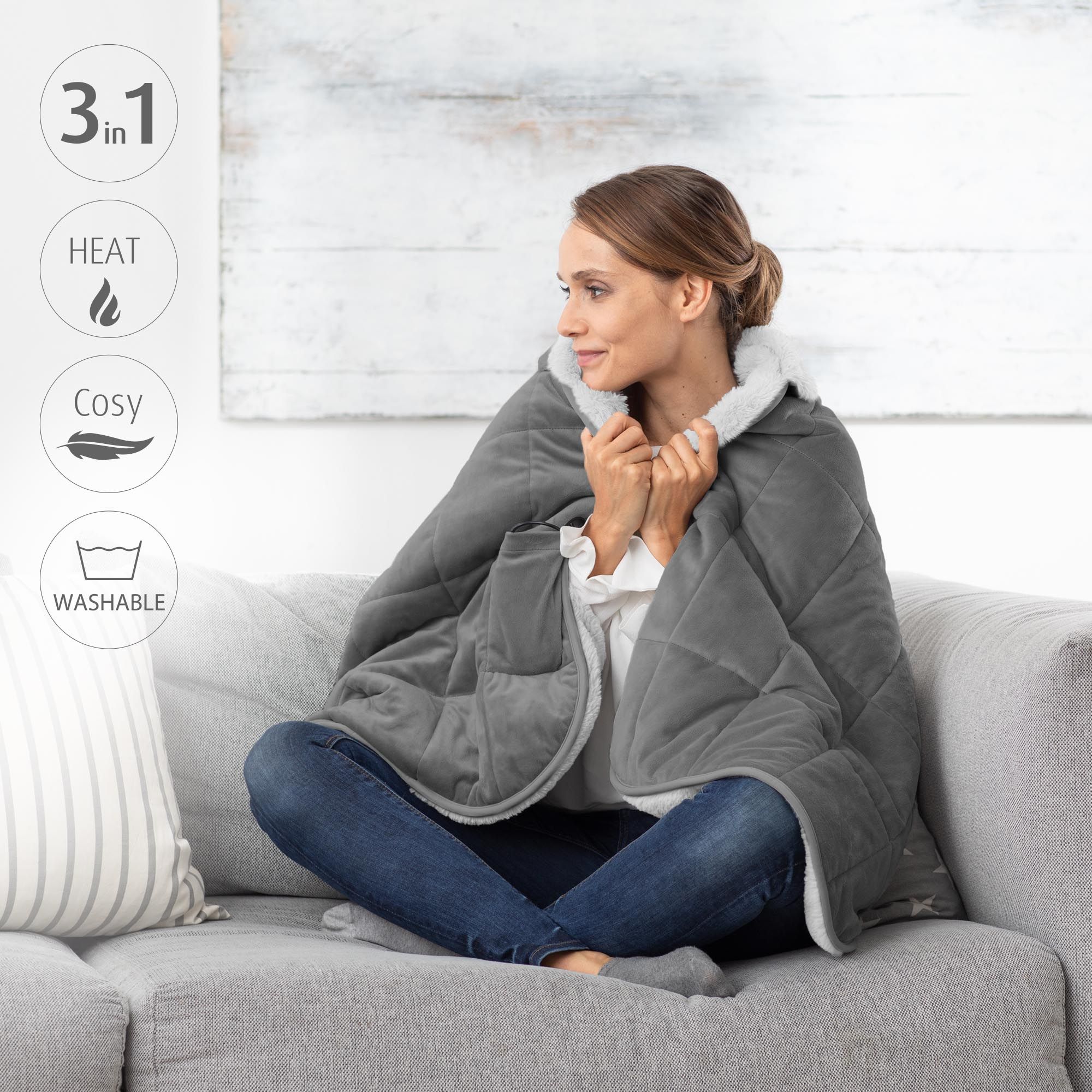 Donna seduta sul divano, indossa una coperta termica grigia. Funzioni 3-in-1: calore, confortevole, lavabile. Simboli.