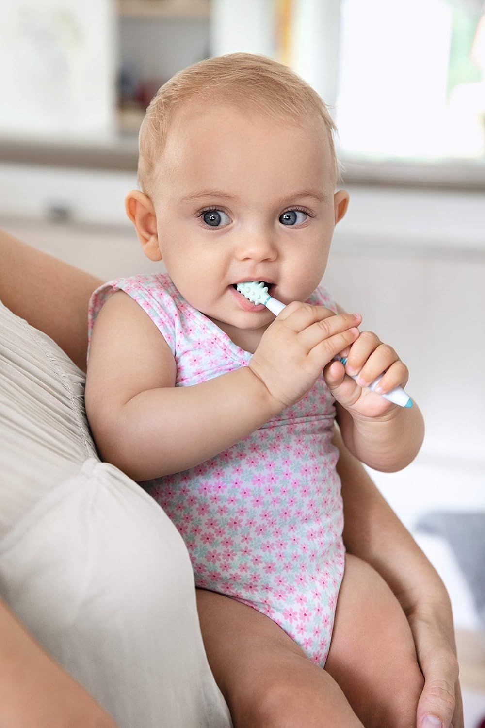Bambino con spazzolino in bocca. Il bambino indossa un body rosa. Il bambino è seduto in grembo a una persona.