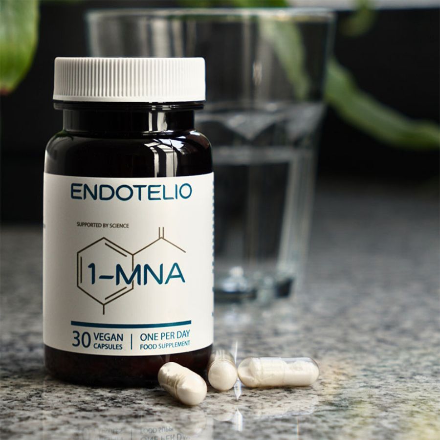 Flacone di vetro marrone con capsule bianche. Etichetta con nome prodotto e 1-MNA. Alcune capsule accanto. Bicchiere d'acqua sullo sfondo.