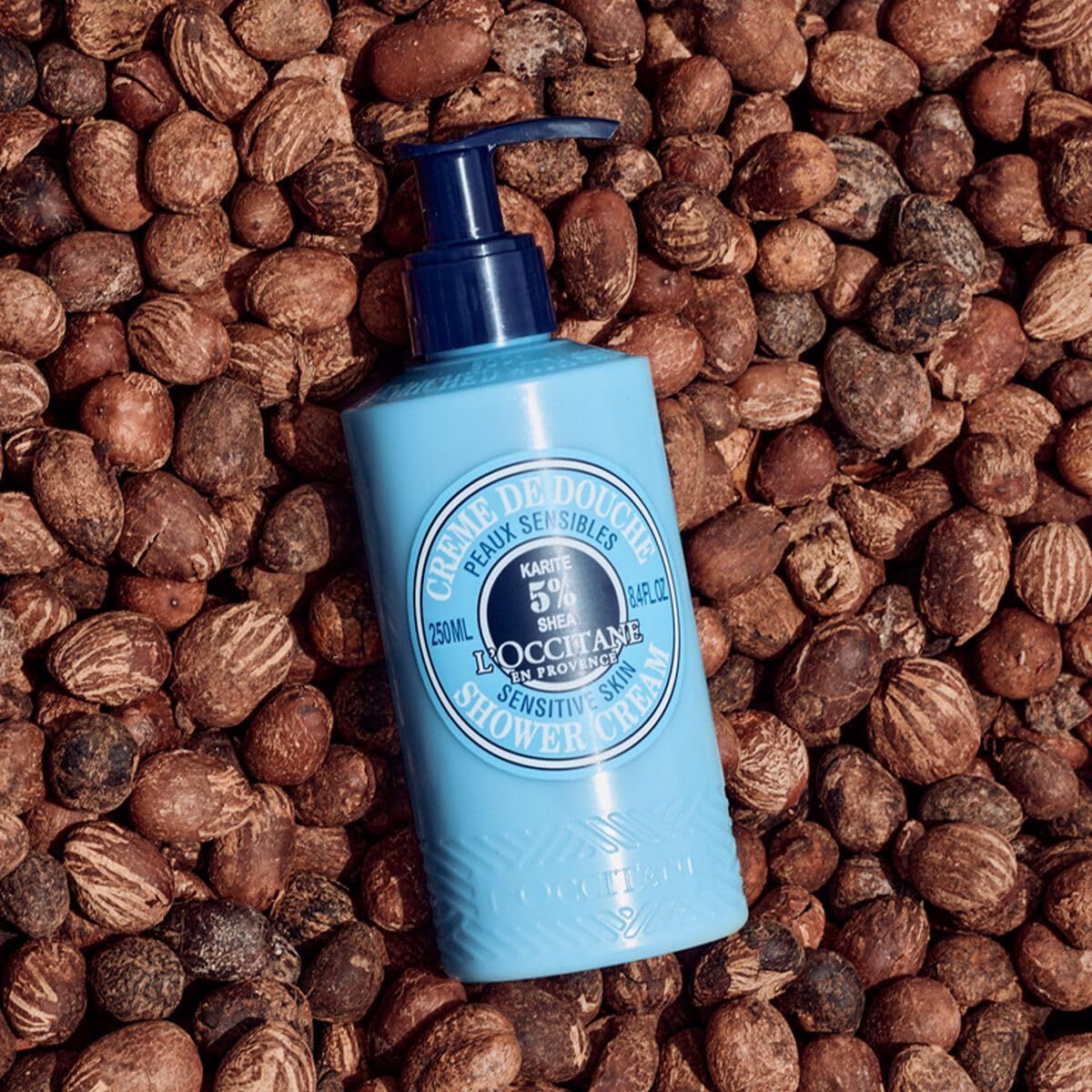 Flacone azzurro con dosatore su noci di karité. Scritta: Crème de Douche, Karité 5% Shea, L'Occitane.